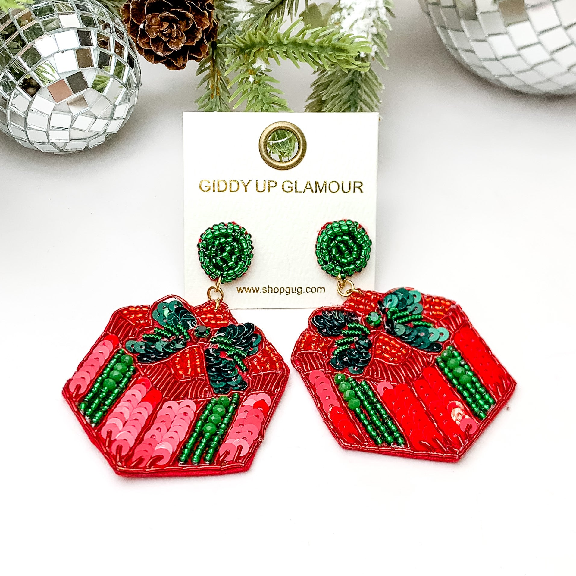 Red and green beaded present shaped earrings. These earrings are pictured on a white background with pine trees at the top of the picture. 