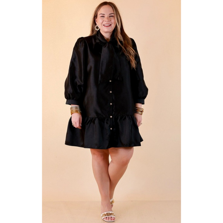 Full length image of a woman wearing the Sealed With a Bow Tie Neck Long Sleeve Button Front Dress with Ruffle Hem in Black
