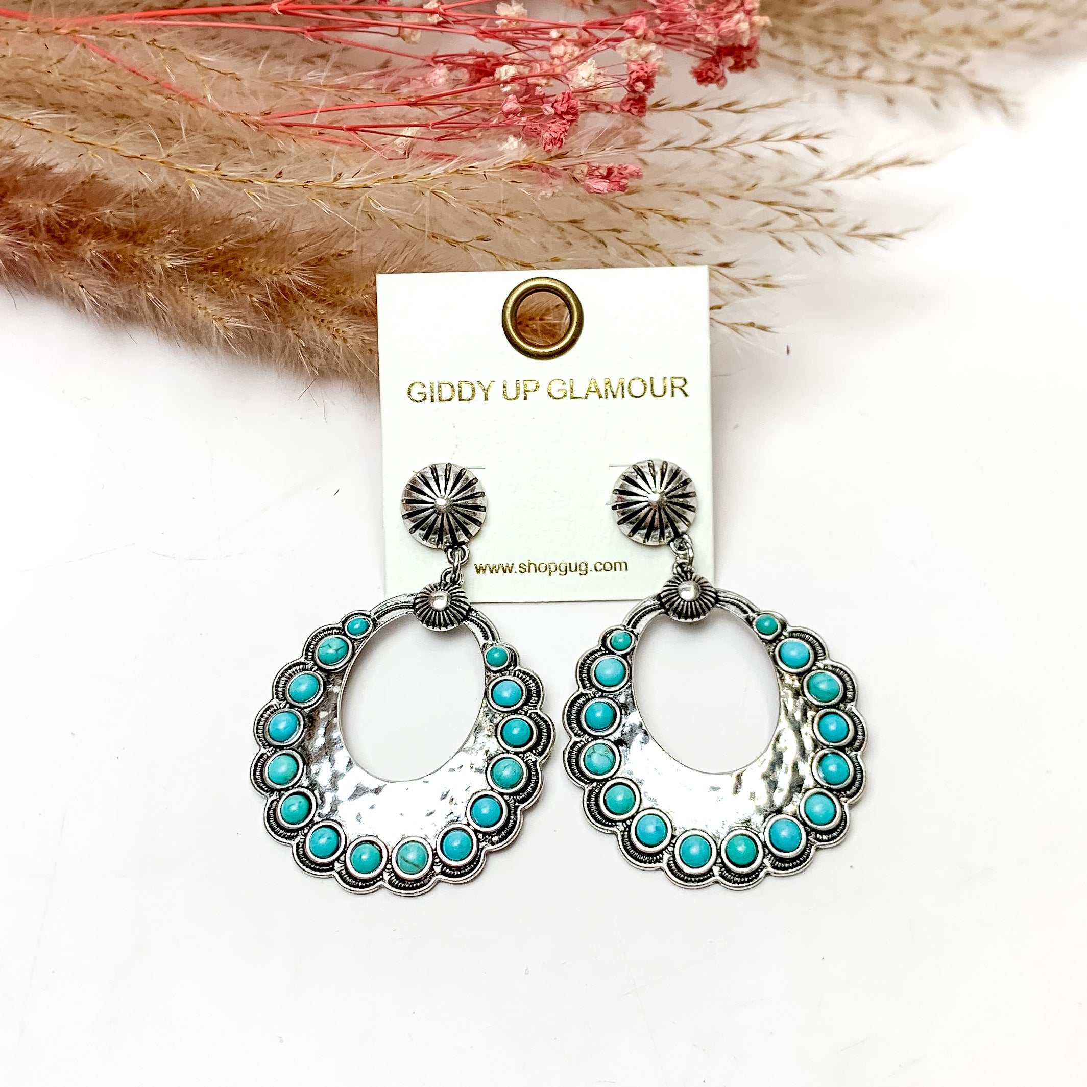Western Open Silver Tone Earrings With Turquoise Stones. These earrings are pictured on a white background with plants behind for decoration.