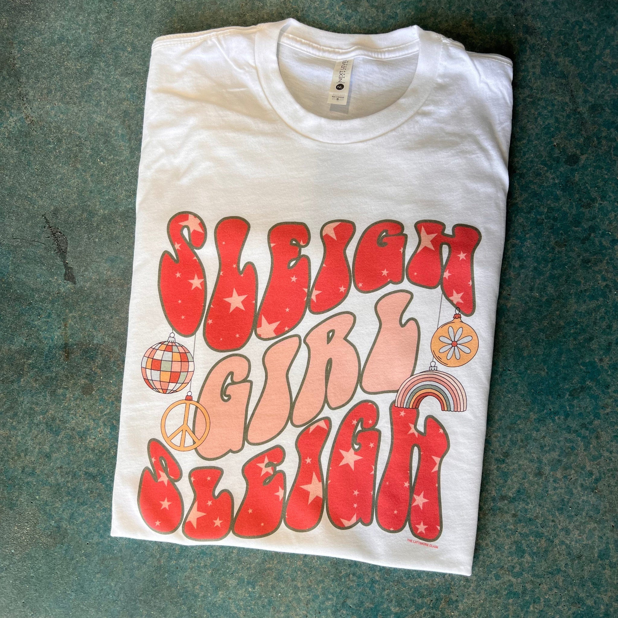 This white tee features a crew neckline, short sleeves, and a hand drawn graphic with the words "Sleigh Girl Sleigh" in a fun, groovy font. The words "Sleigh" is red with light pink stars throughout, and the word "Girl" is light pink".  There are four different ornaments hanging from the first "Sleigh" that are a disco ball, peace sign, rainbow, and a floral ornament. These are in fun, light colors and this graphic tee is shown here as a folded flatlay. 