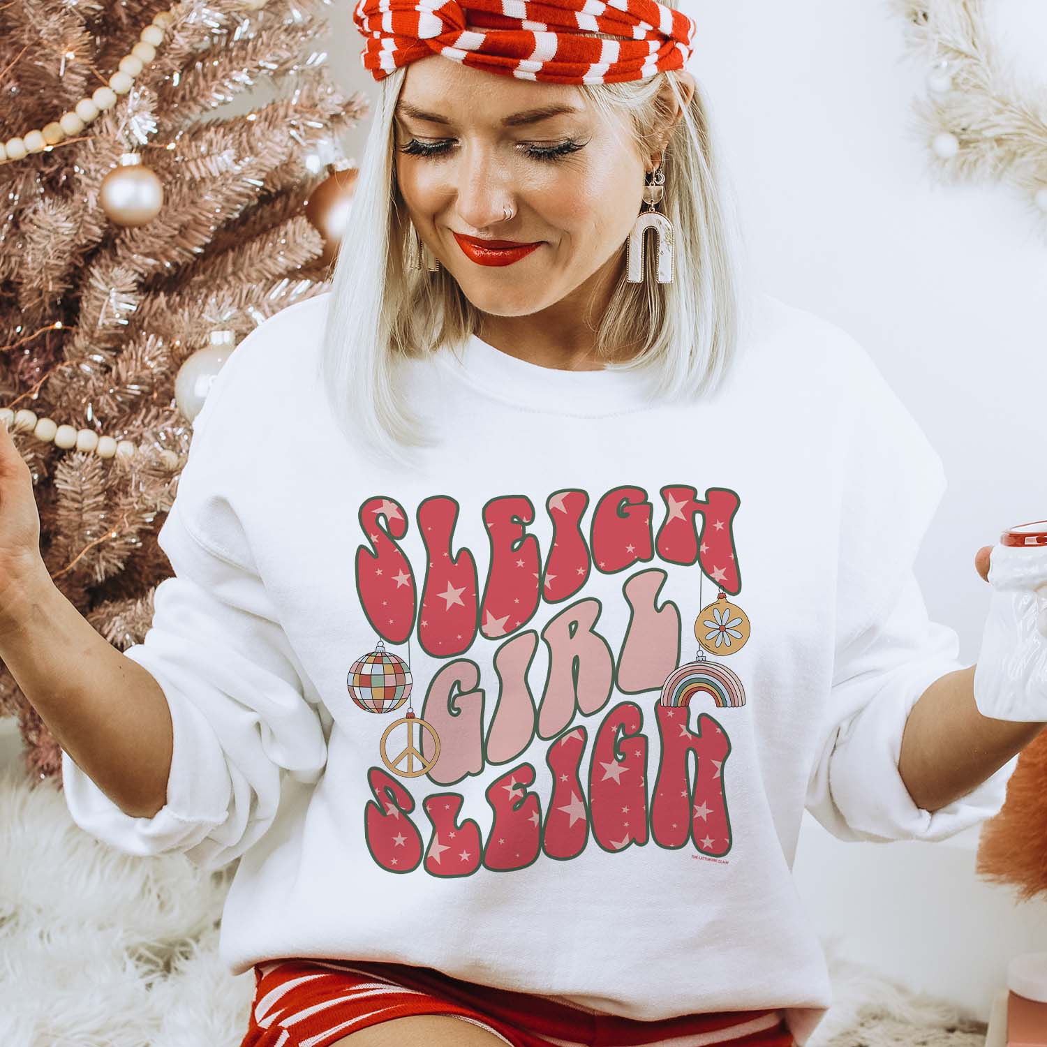 This white sweatshirt features a crew neckline, long sleeves, and a hand drawn graphic with the words "Sleigh Girl Sleigh" in a fun, groovy font. The words "Sleigh" is red with light pink stars throughout, and the word "Girl" is light pink". There are four different ornaments hanging from the first "Sleigh" that are a disco ball, peace sign, rainbow, and a floral ornament. These are in fun, light colors.