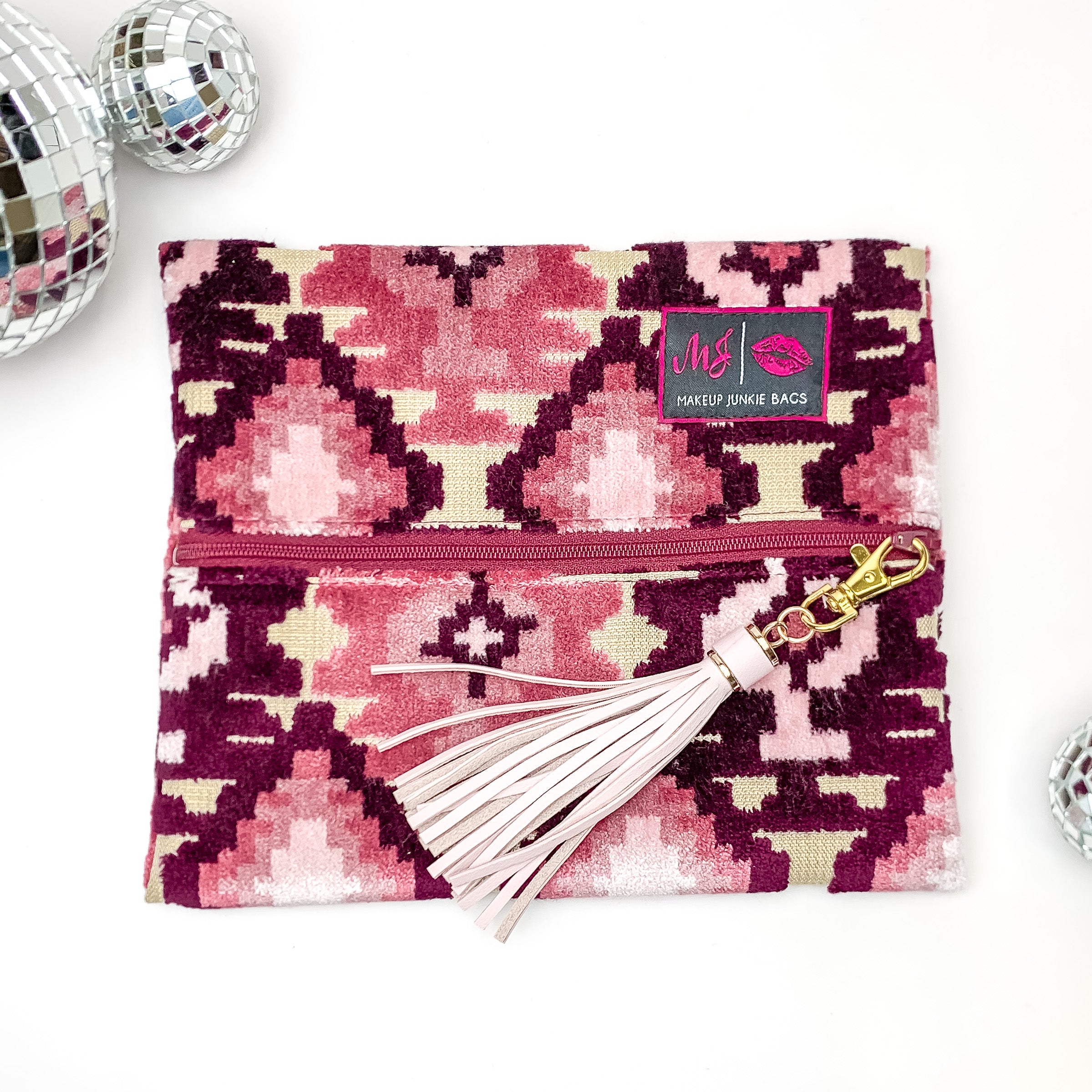 Pictured on a white background with disco balls at top is a mini lay flat bag in a maroon aztec print. This bag includes a middle zipper and a tassel. 