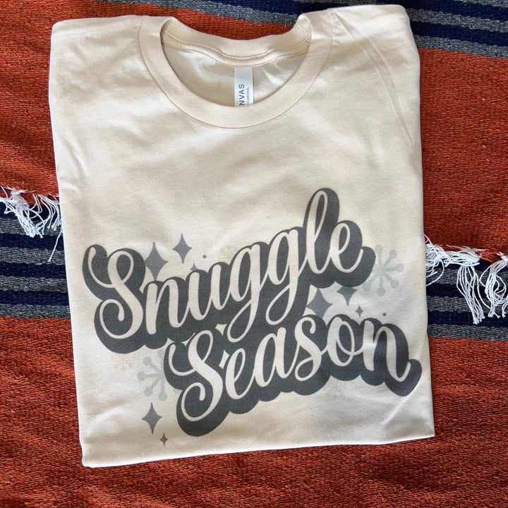 This cream tee includes a crew neckline, short sleeves, and a graphic that says "Snuggle Season" in a cute, cursive font in white with a black outline. There are also a few diamonds and snowflakes around the words. This is pictured as a folded flatlay on a serape background. 