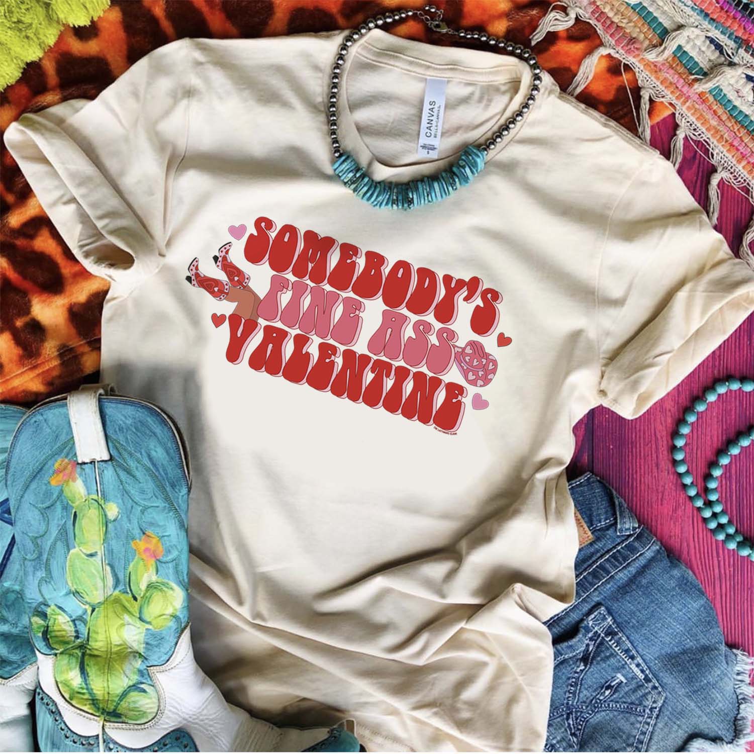 This crew neck cream tee is shown here as a flatlay with rolled sleeves on a multi-colored background. It is paired with a turquoise necklace, denim shorts, and turquoise boots with a cactus. The graphic says "Somebody's Fine Ass Valentine" in pink and red bubble font with hearts throughout. There is also legs with cowboy boots on the left side (looking at the tee) and a pink cowboy hat with hearts on the right side of the graphic (if looking directly at the tee). 