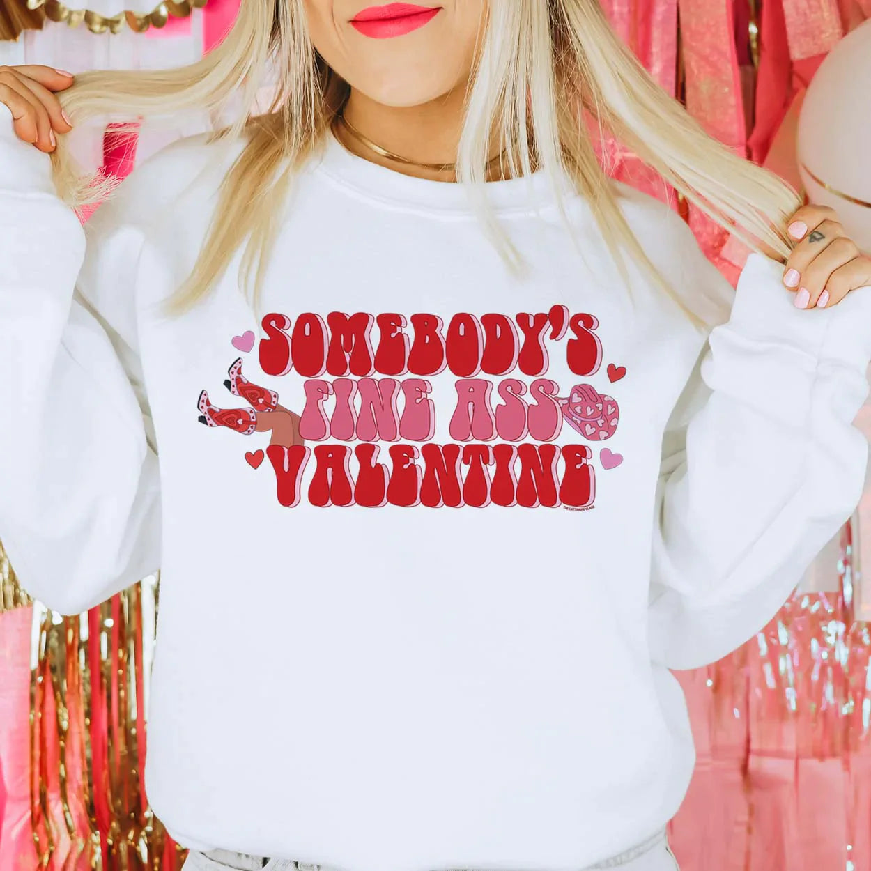 This crew neck white sweatshirt is shown here modeled with jeans and a gold necklace. The graphic says "Somebody's Fine Ass Valentine" in pink and red bubble font with hearts throughout. There is also legs with cowboy boots on the left side (looking at the tee) and a pink cowboy hat with hearts on the right side of the graphic (if looking directly at the tee).