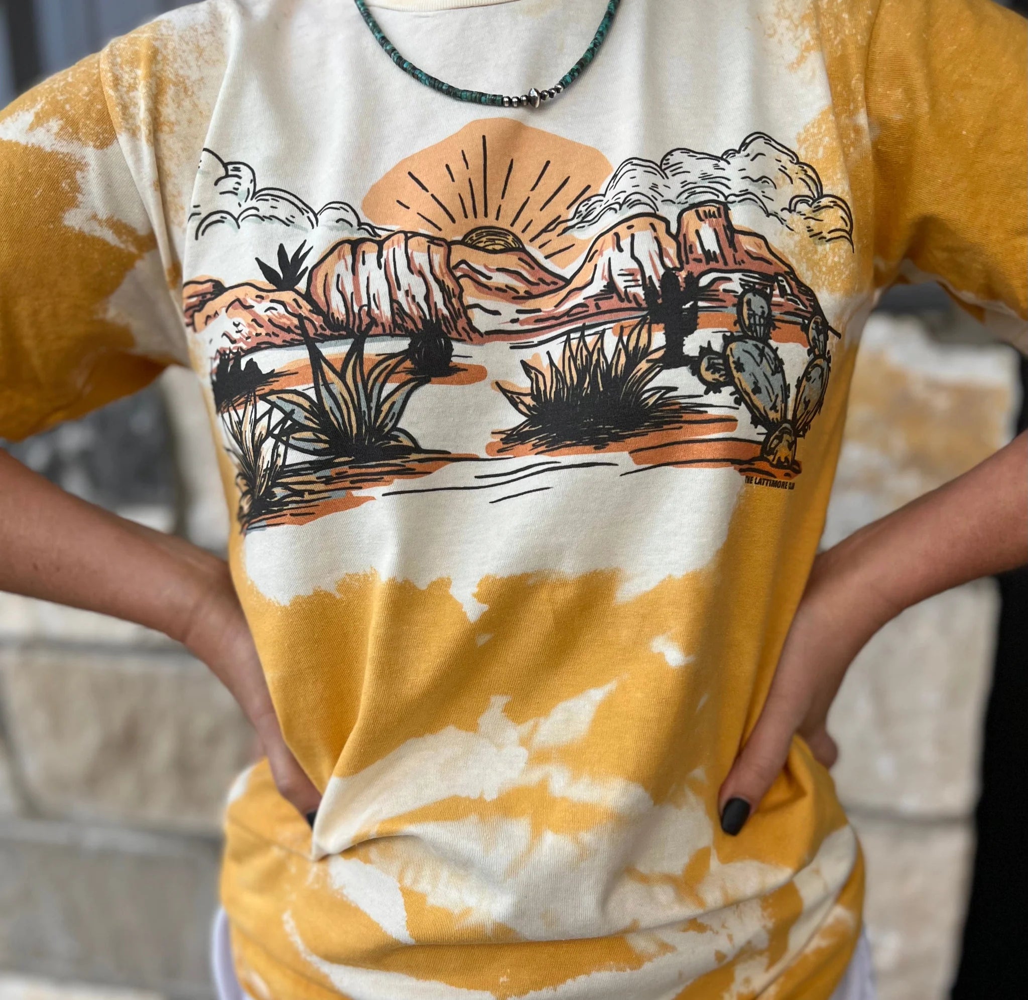 A bleached patterned mustard color short sleeve crewneck tee with a graphic in the center featuring cacti, desert flora, cliffs, and a sunset. Item is pictured on a light brick background