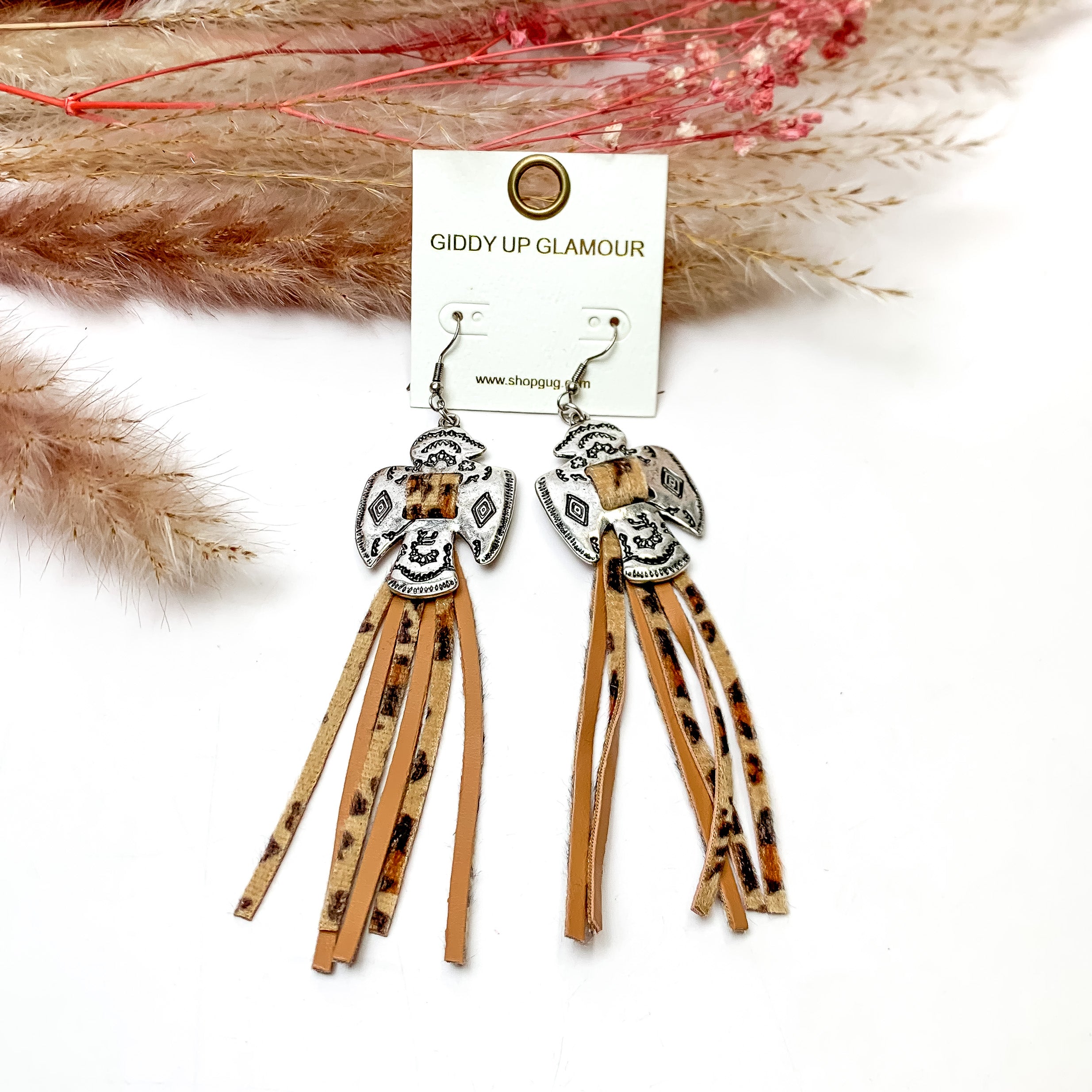 Thunderbird Tassel Earrings in Leopard. These earrings are pictured on a white background with flowers behind for decoration.