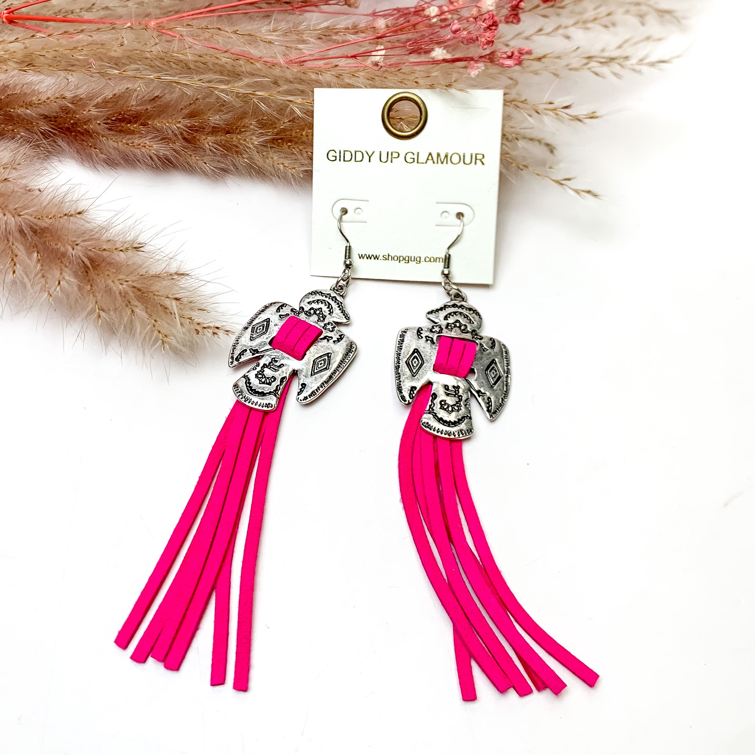 Thunderbird Tassel Earrings in Fuchsia. These earrings are pictured on a white background with flowers behind for decoration.