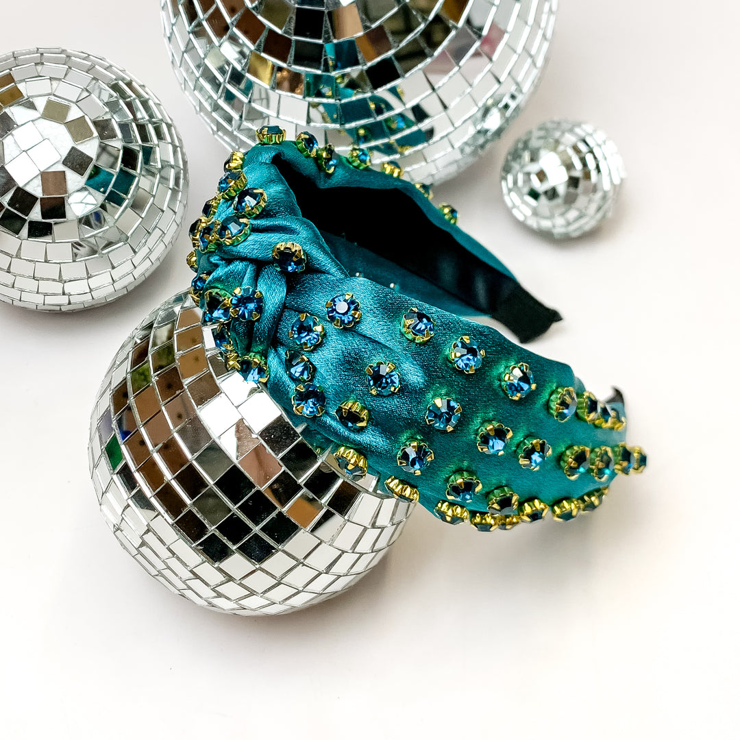 Crystal Detailed Satin Knot Headband in Teal. This headband is pictured on a white background and is surrounded by disco balls.