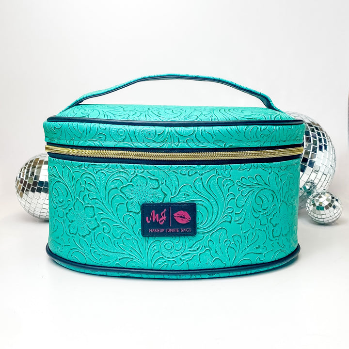 Pictured on a white background with disco balls in the background is a traincase with a top handle in a turquoise leather tooled print. This bag includes a middle zipper and a tassel.