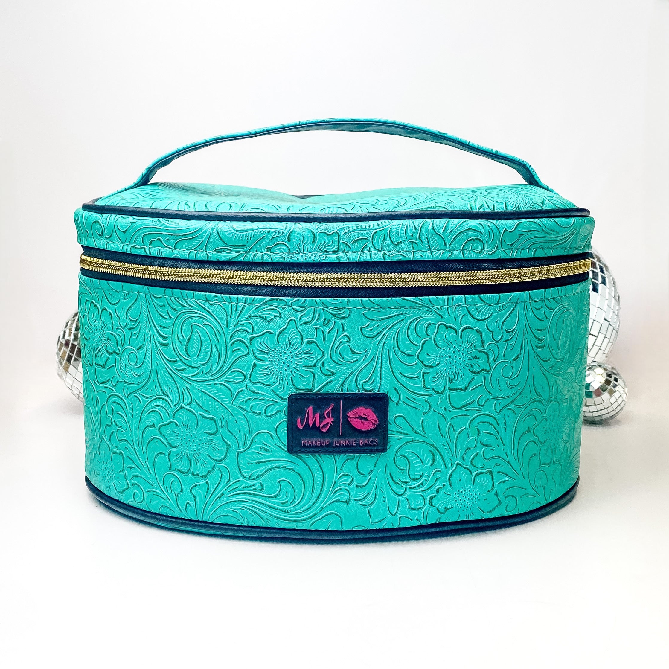 Pictured on a white background with disco balls in the background is a traincase with a top handle in a turquoise leather tooled print. This bag includes a middle zipper and a tassel.