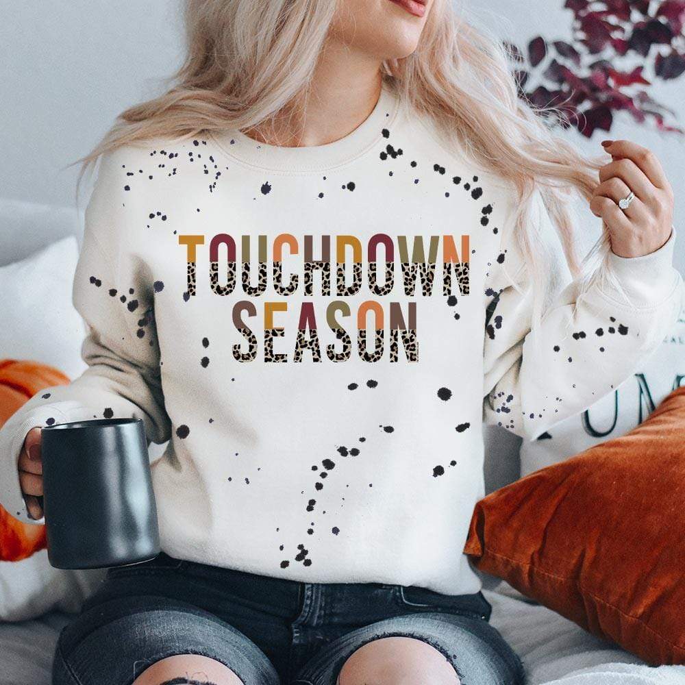 Model is wearing a white sweatshirt featuring a black splatter print and a half multicolor half leopard print graphic that says "Touchdown season"