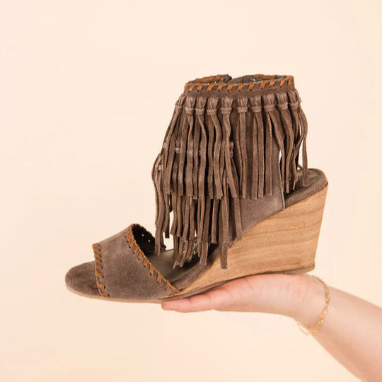 A solid background with a hand holding a taupe wedge with fringe coming from the ankle strap and a toe strap.