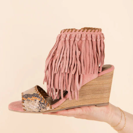 A solid cream color background with a hand holding a blush pink wedge shoe that has fringe and a snake inspired toe strap.
