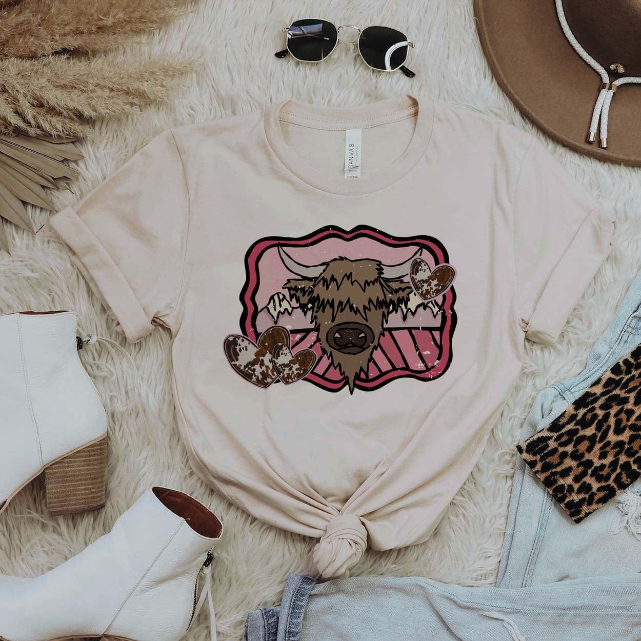 An ivory tee shirt that is knotted in the front and sleeves cuffed. Tee shirt has a highlander cow head drawn on a mountain background with pink detailing and cow print hearts. Tee shirt is pictured on fur background with white booties, light wash jeans, sunglasses, and a brown hat.
