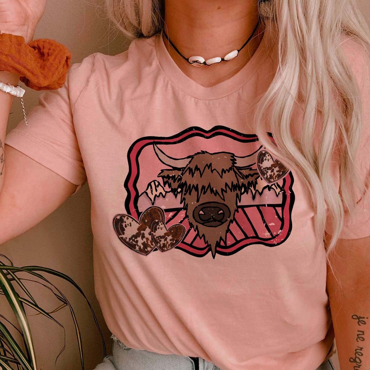 Model is wearing a rose pink tee shirt that has a highlander cow graphic on a mountain background with pink detailing and cowprint hearts. Model has it paired with a shell necklace, a rust scrunchie, and light wash jeans.