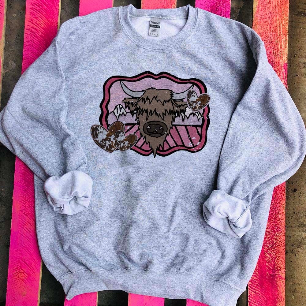 This gray sweatshirt is shown here as a flatlay on a pink wooden background. A highlander cow graphic on a mountain background with pink detailing and cowprint hearts. It is shown with the sleeves rolled as well. 