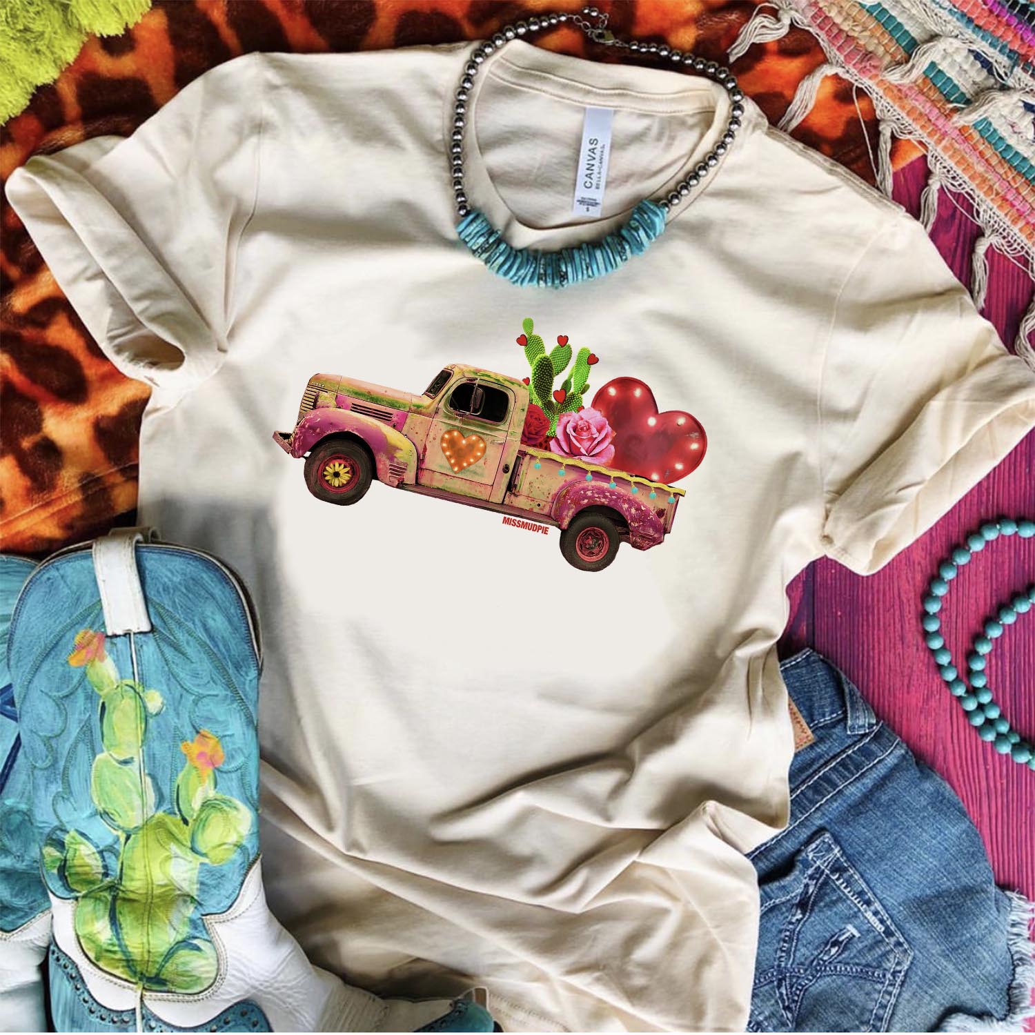 An ivory tee shirt laid on a purple wooden background with cowgirl boots, denim shorts, turquoise and silver necklace, and mix print rugs. The tee shirt has a graphic that is a rustic truck with a cactus, heart, and rose in the bed of the truck.