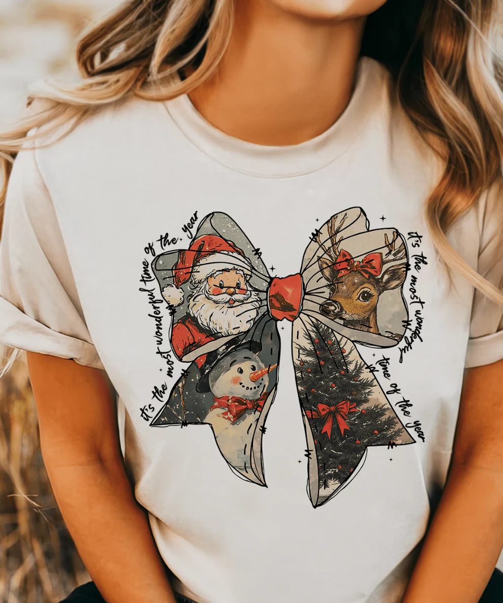 White t-shirt with festive design featuring Santa Claus, a mouse, and a snowman on a blurred natural background.