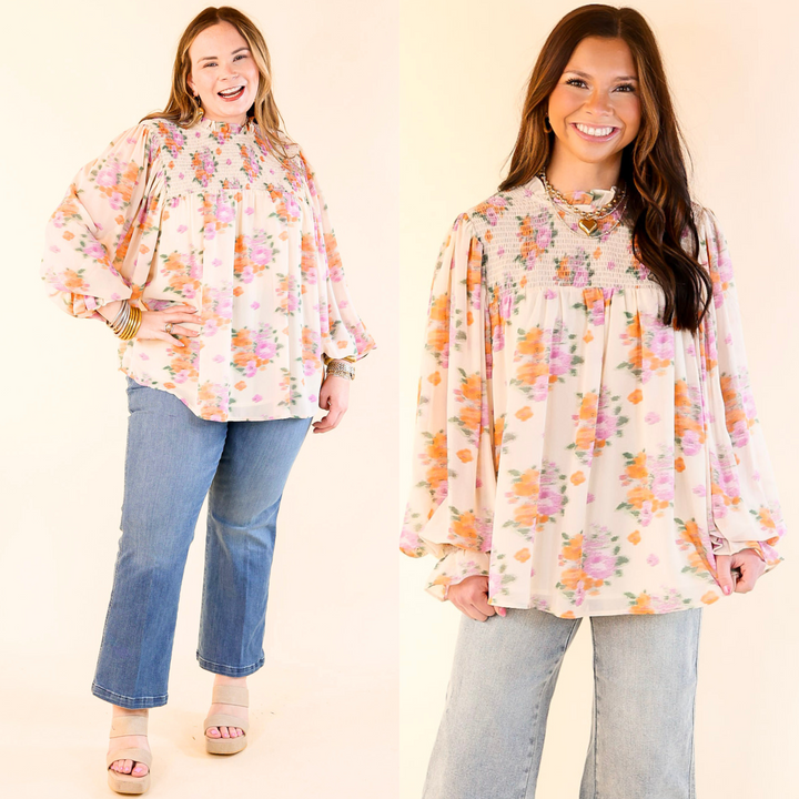 Top includes long sleeves with a ruffle wrist, a high ruffle neckline, a smocked upper, and a watercolor floral print that is green, orange, and purple. 