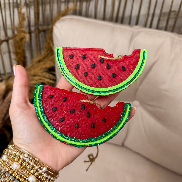 A rustic wooden background with a hand holding 2 watermelon shaped freshies