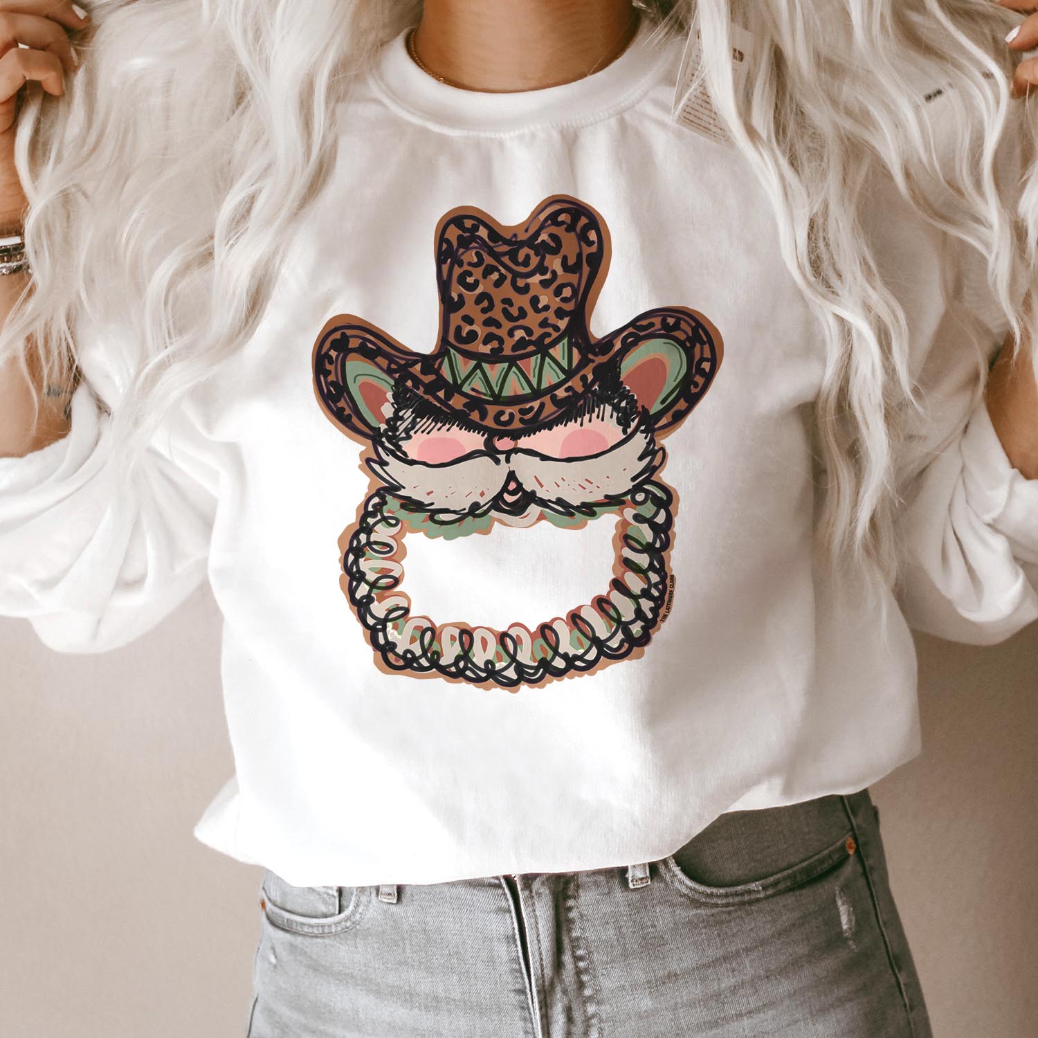 This white sweatshirt includes a crew neckline, long sleeves, and a Santa Clause graphic with a leopard print hat. The model has this sweatshirt styled with rolled sleeves and light wash denim jeans. 