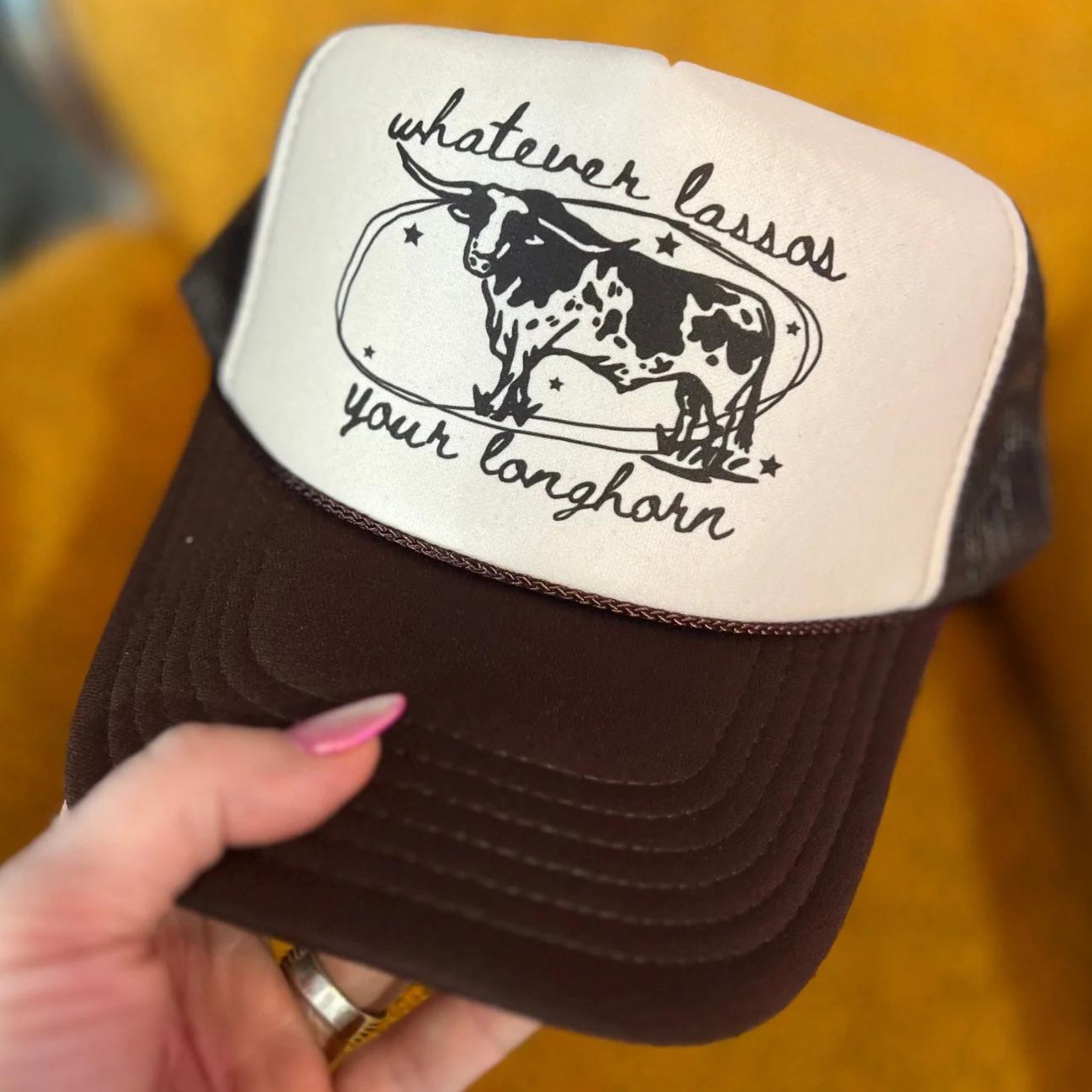 Photo features a brown and cream trucker hat with the words "whatever lassos your longhorn" with a picture of longhorn on the front.