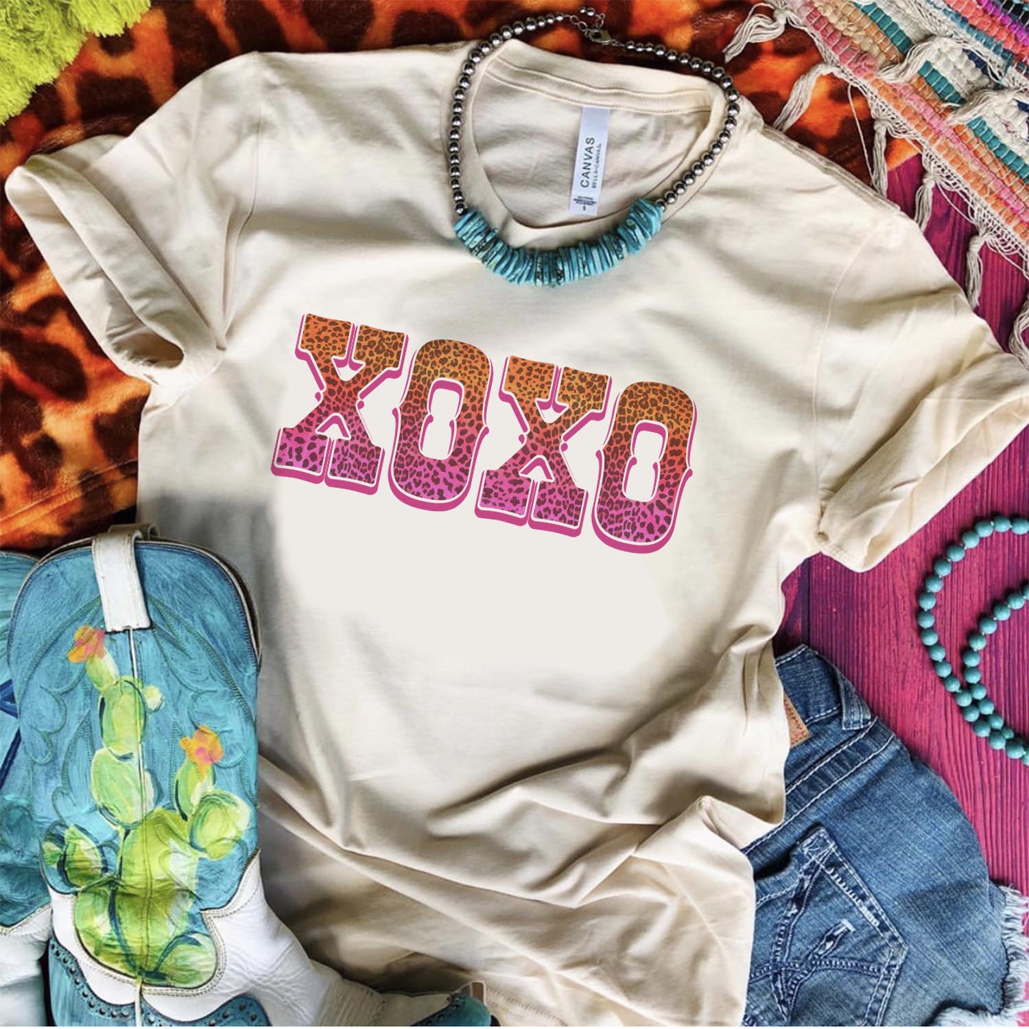 An ivory tee shirt laid on a purple wooden background with cowgirl boots, denim shorts, turquoise and silver necklace, and mix print rugs. The tee shirt has a graphic that says "XOXO" in an ombre leopard print.