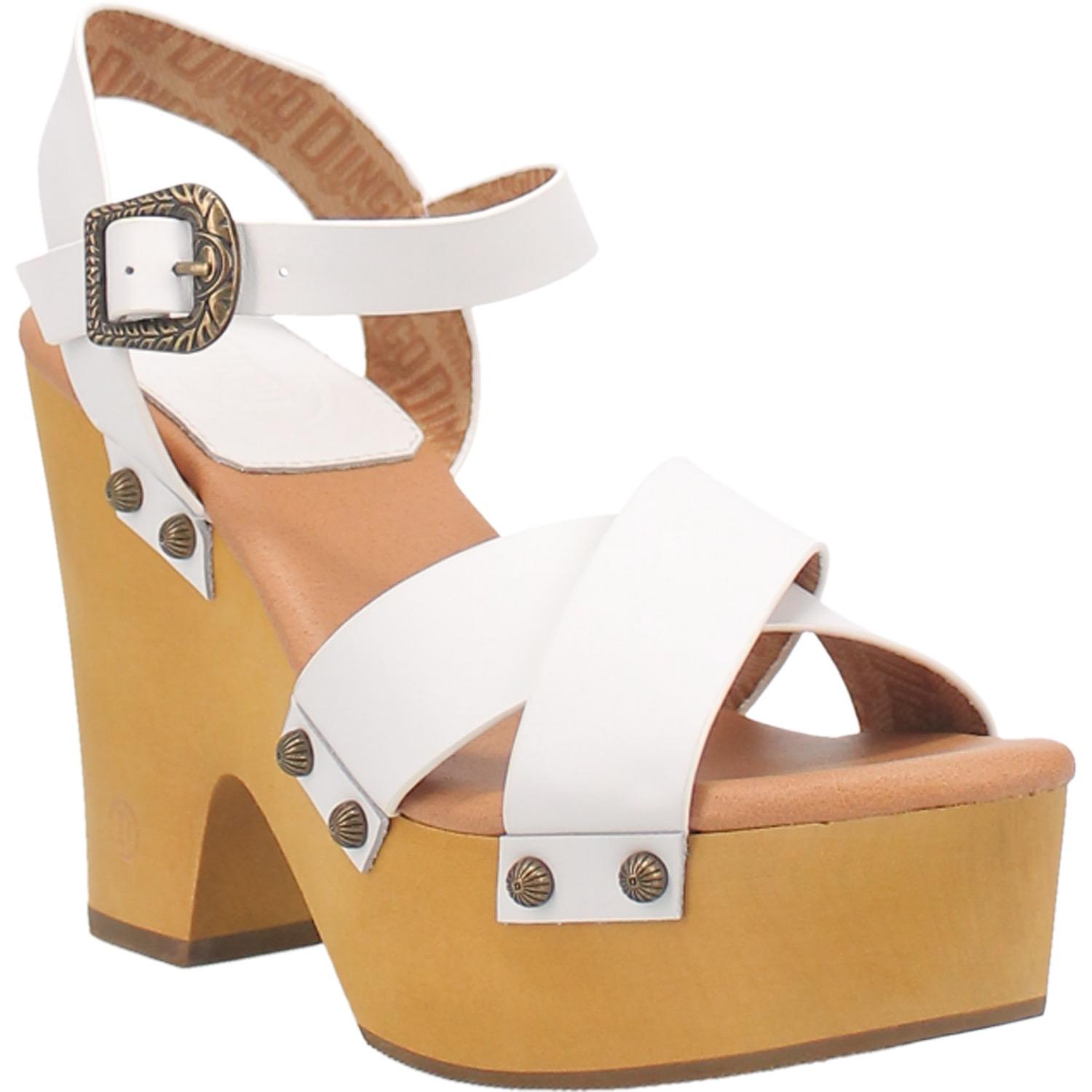 A platform high heeled white sandal with two crossed whited straps in the front, white ankle straps with a unique buckle, and circular studs. Item is pictured on a white background.