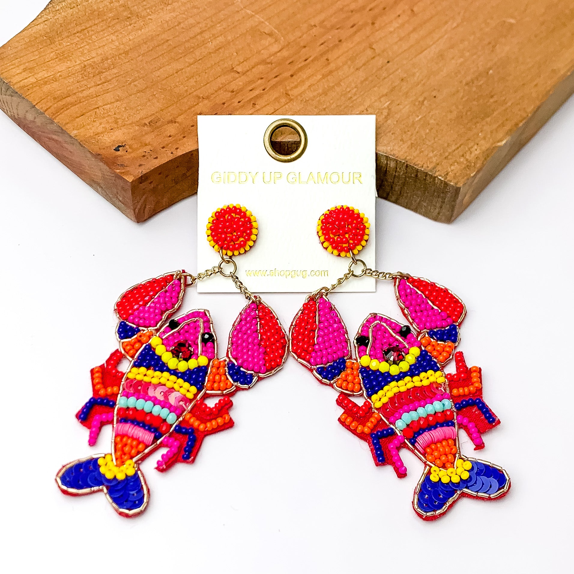 Pictured are beaded lobster earrings with multi beaded colors with gold outline detailing. These earrings are propped up on a piece of wood with a white background.