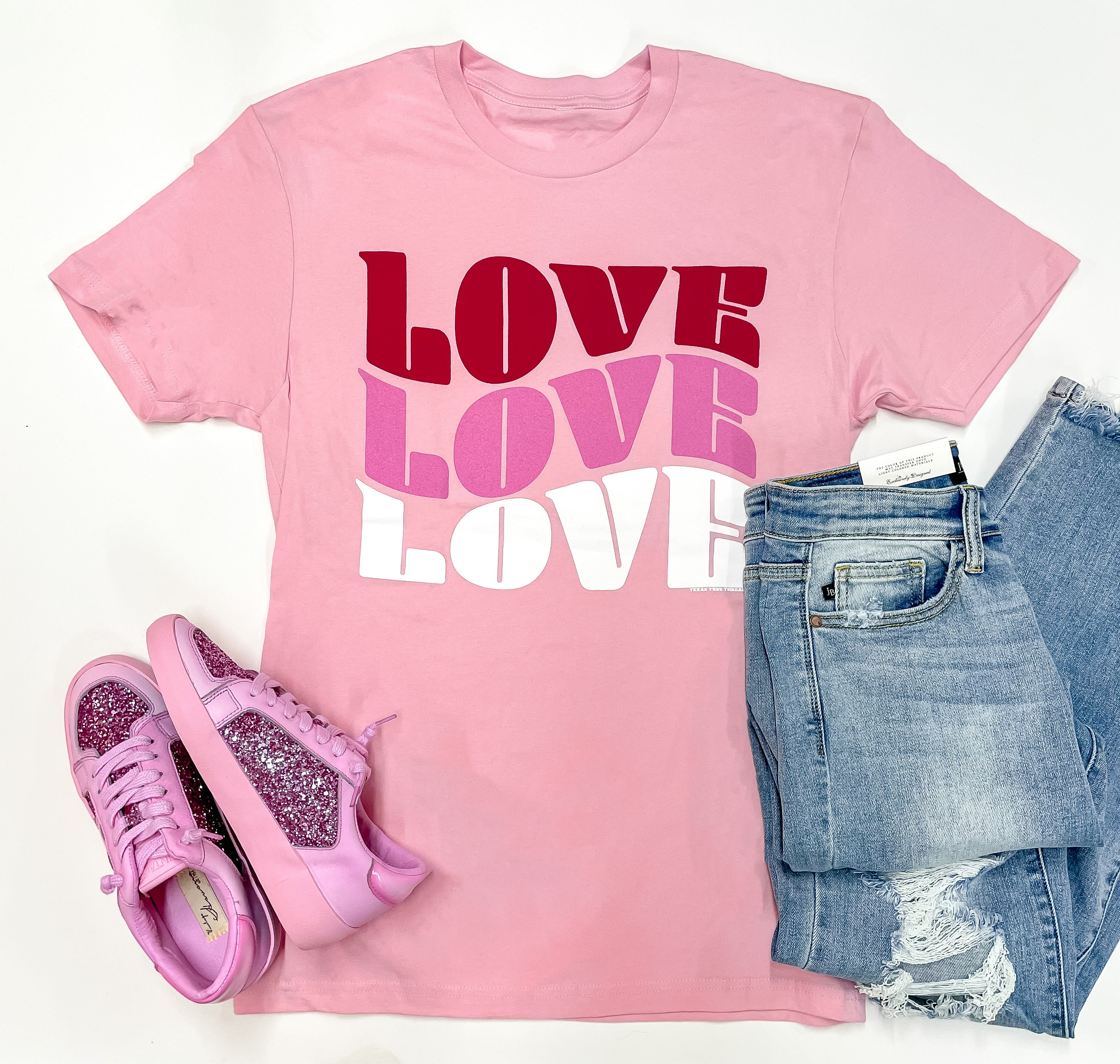 A light pink tee shirt with a graphic that says "LOVE" three times, in red, hot pink, and white. This tee shirt is pictured on a white background with distressed jeans and hot pink sneakers to match.