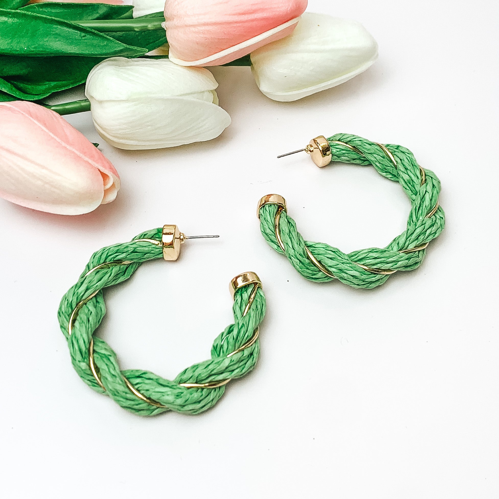 Pictured are light green raffia twisted hoop earrings with gold detailing.  They are pictured with pink and white tulips on a white background.