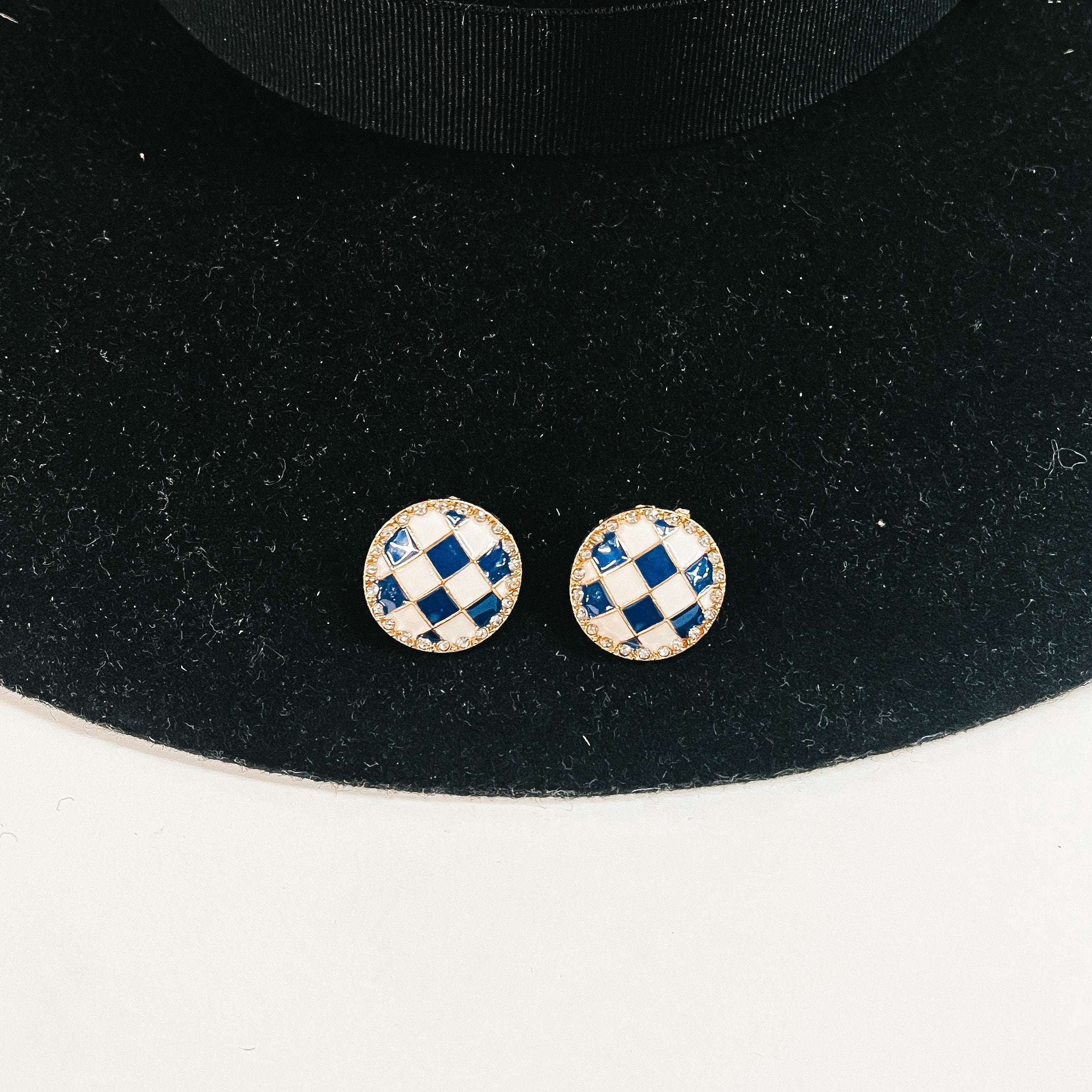 This is a pair of checkered patterned stud earrings in white/blue in a gold  setting with clear crystals all around. This pair of earrings is laying on a  black felt hat brim and on a white background.
