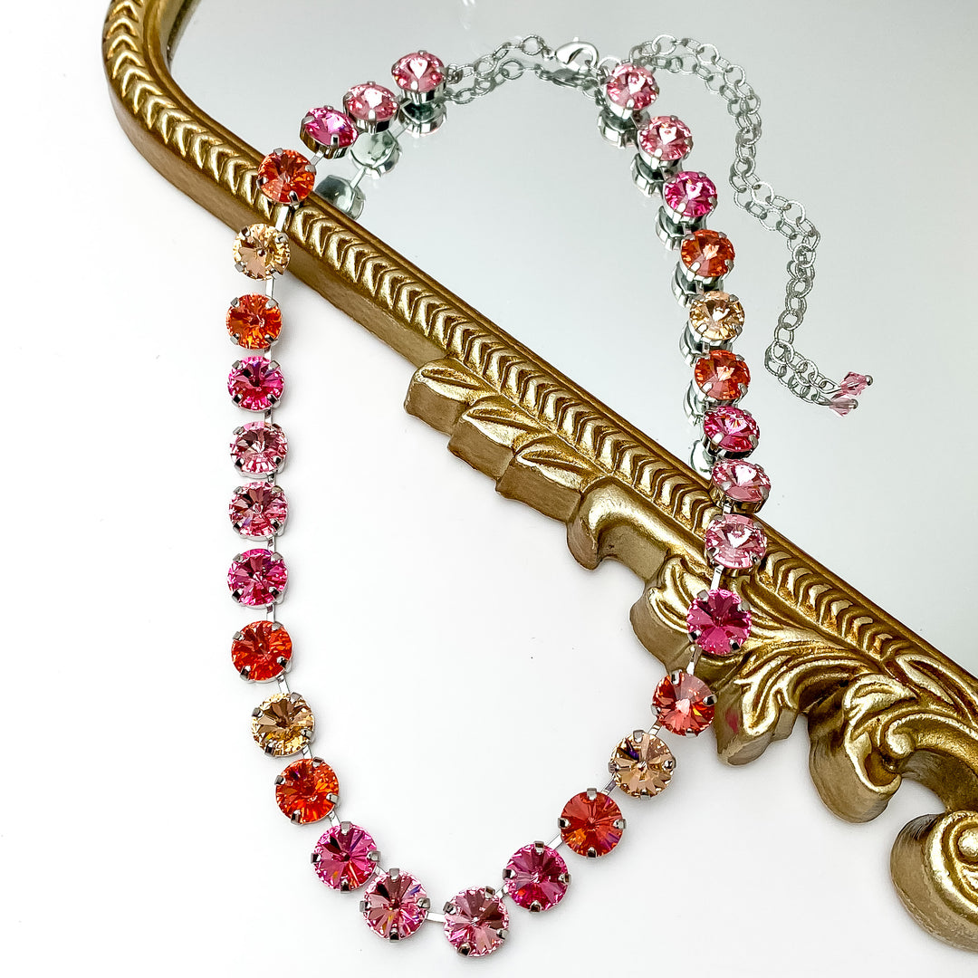 Mix of pink round crystal necklace. This necklace has a silver setting. This necklace is pictured partially laying on a gold mirror on a white background.    