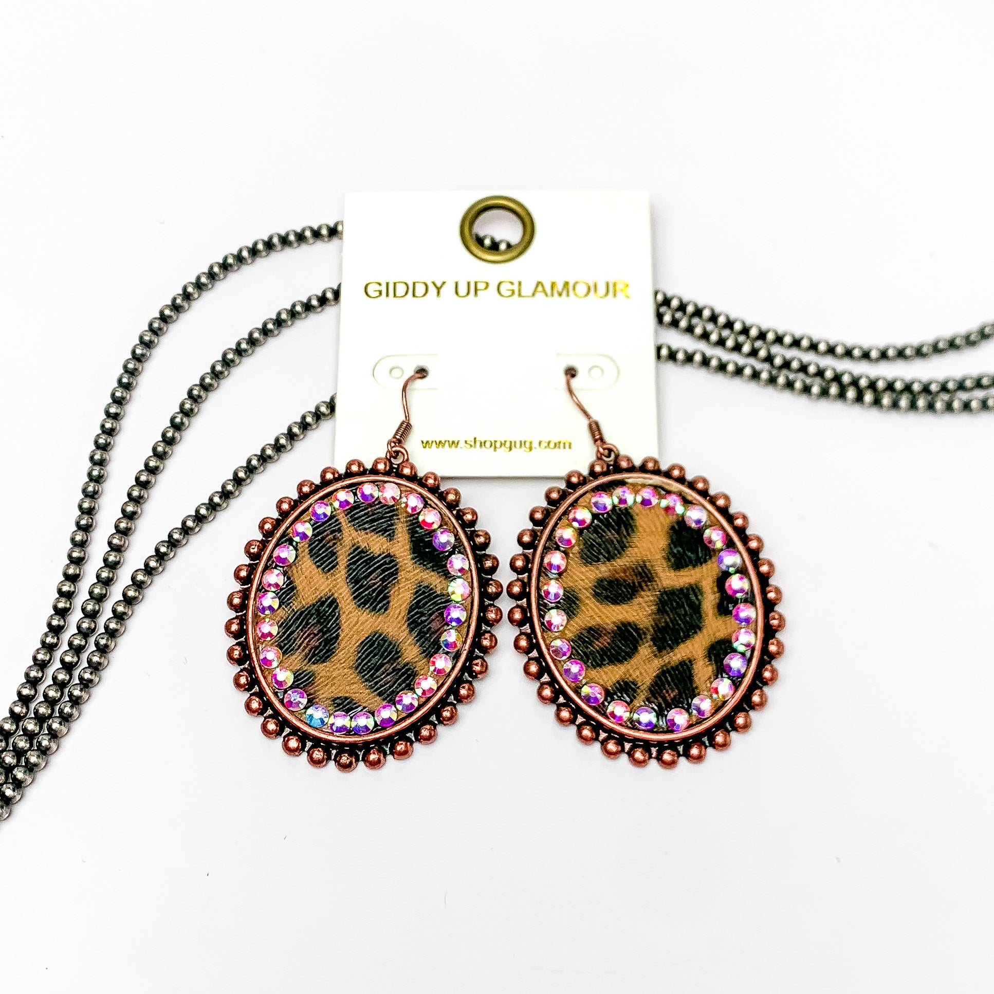 These earrings are oval shaped with leopard print inlay with the outline of the earring is copper but also has ab crystal going around the oval shape. These pair of earrings are pictured on a white background with navajo pearls behind it.