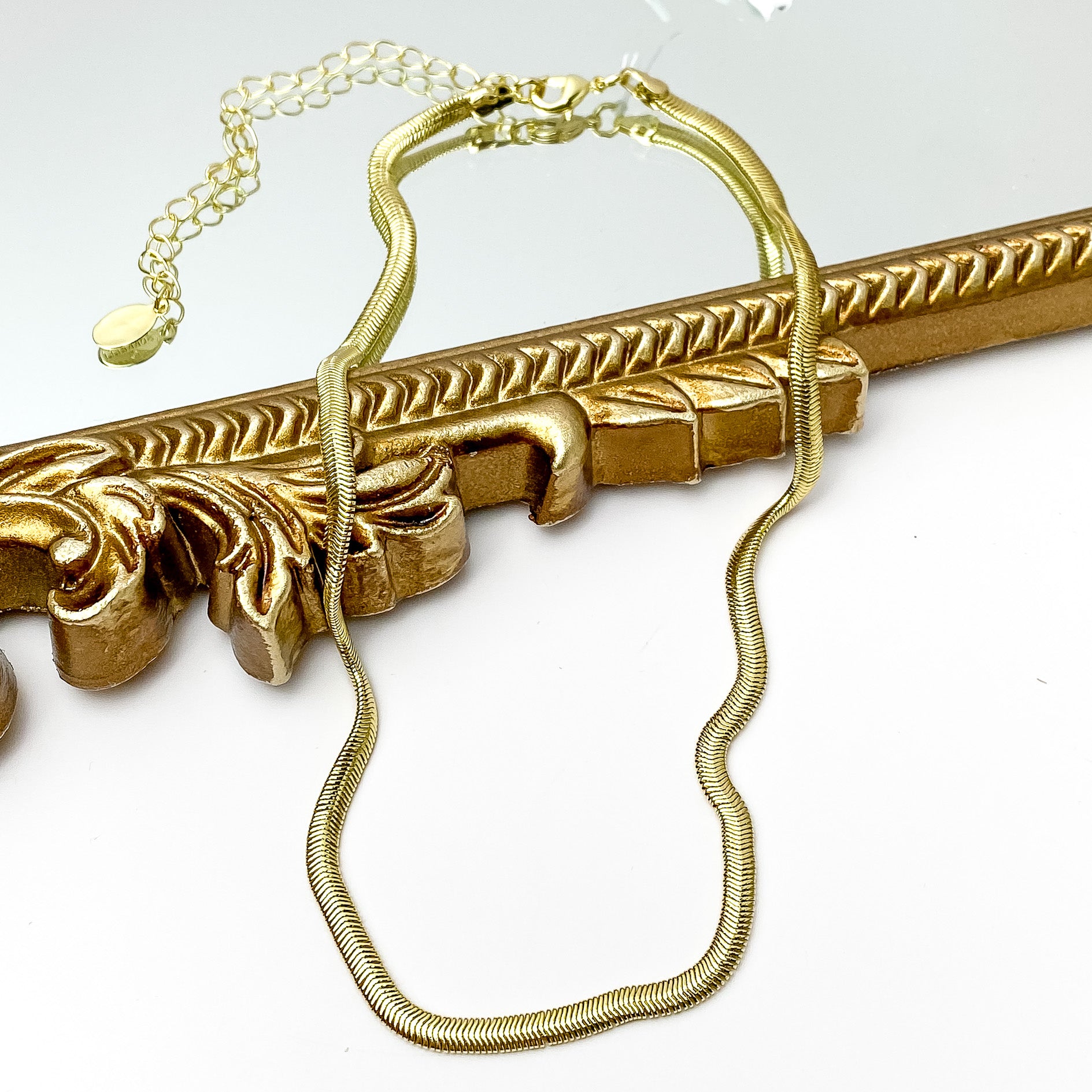 Gold herringbone chain necklace. This necklace is pictured laying partially on a gold mirror on a white background.   