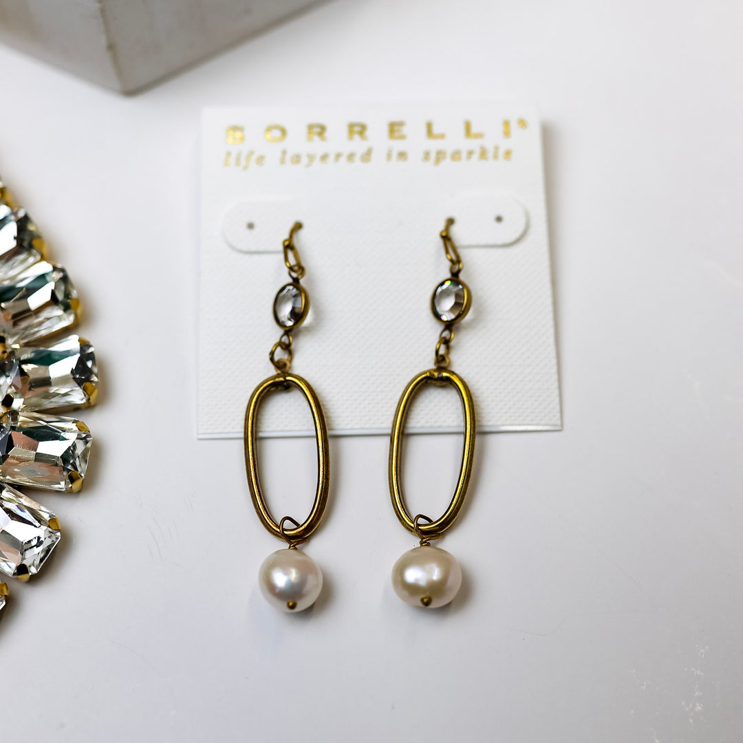 A pair of gold tone oval outline earrings with a pearl pendant at the bottom. Pictured on a white background with crystal necklaces.