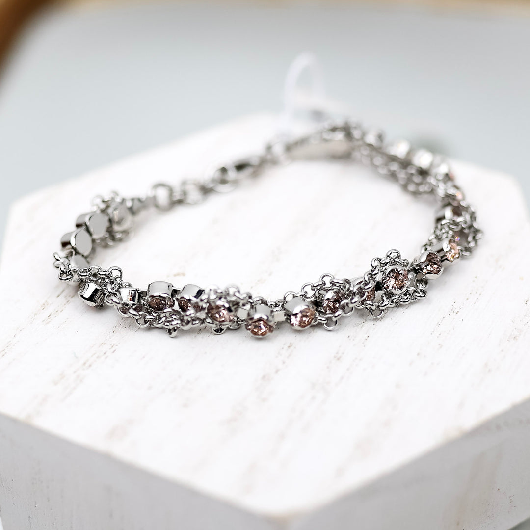 A silver-tone tennis bracelet with blush pink crystals and chain detailing. Pictured on a white background.