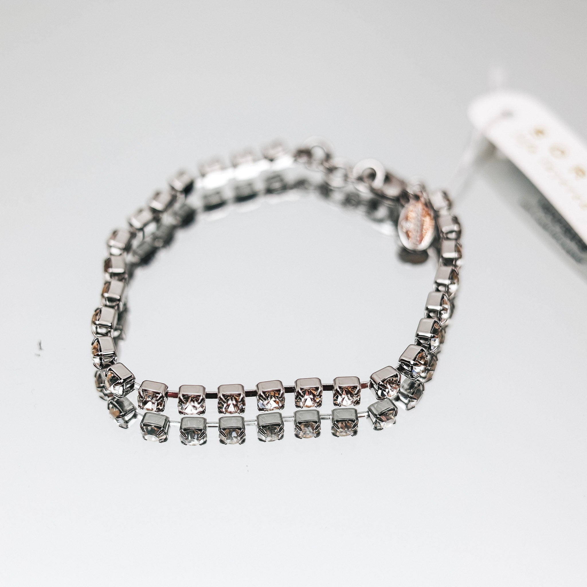 A silver-tone tennis bracelet with small clear square cut crystals pictured on a mirror.