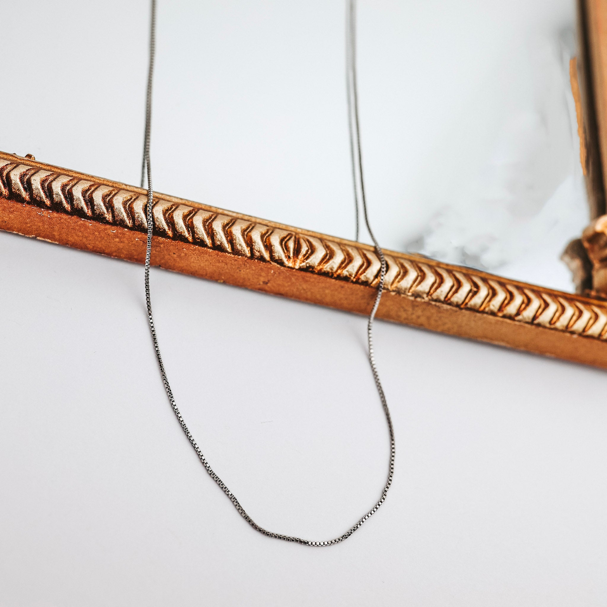 A silver-tone thin box chain necklace that is pictured on a white background with a mirror.