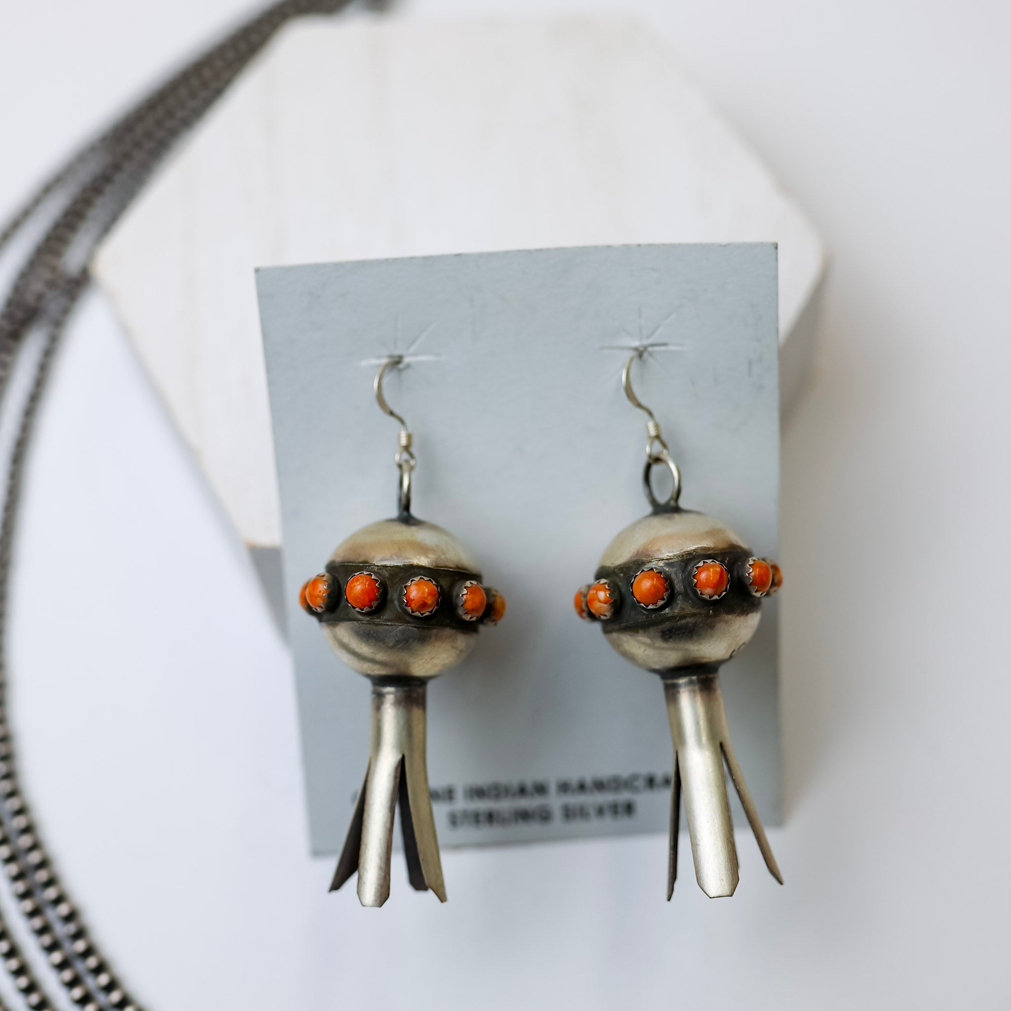 Monica Smith Navajo Handmade Sterling Silver Blossom Earrings with Orange Spiny Oyster is centered in the picture. Navajo pearls are laid to the left of the picture. All on a white background. 