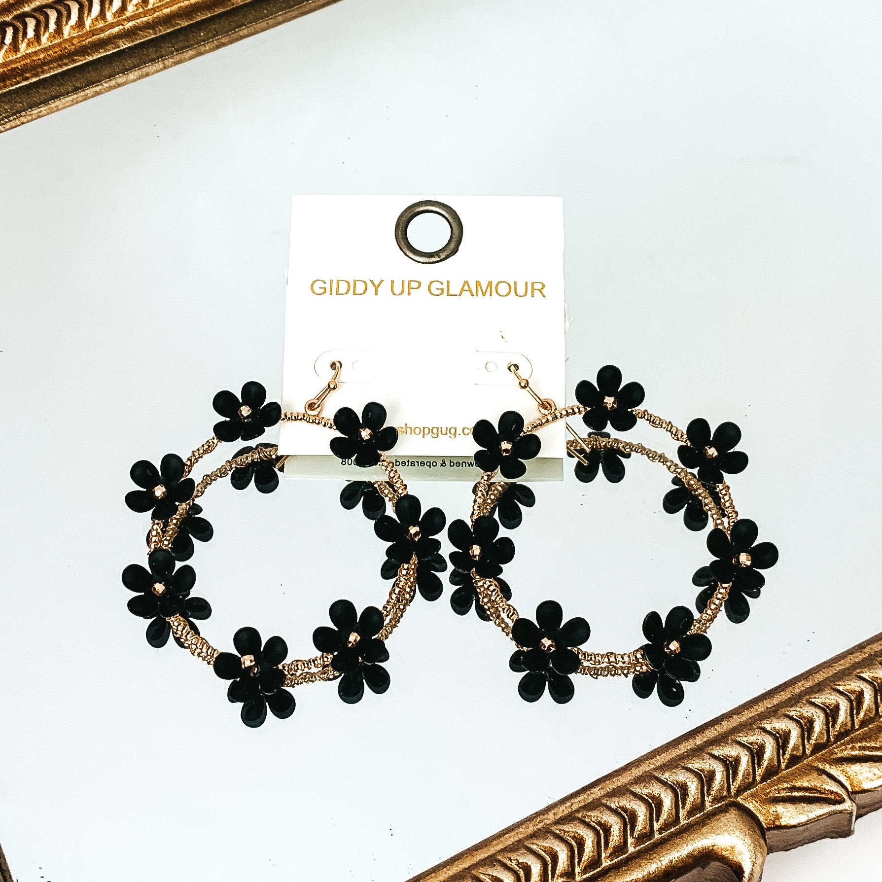 Gold, open circle drop earrings with black flower charms outlining the earrings. These flowers have a gold bead center. These earrings are pictured laying on a gold mirror on the white background. 