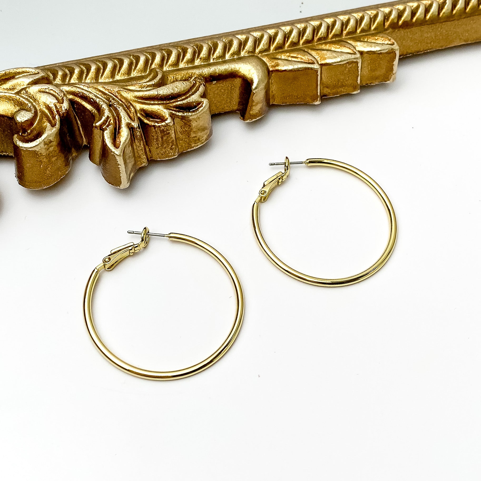 Pictured are a pair of gold hoop earrings. These earrings are pictured in front of a gold mirror on a white background. 