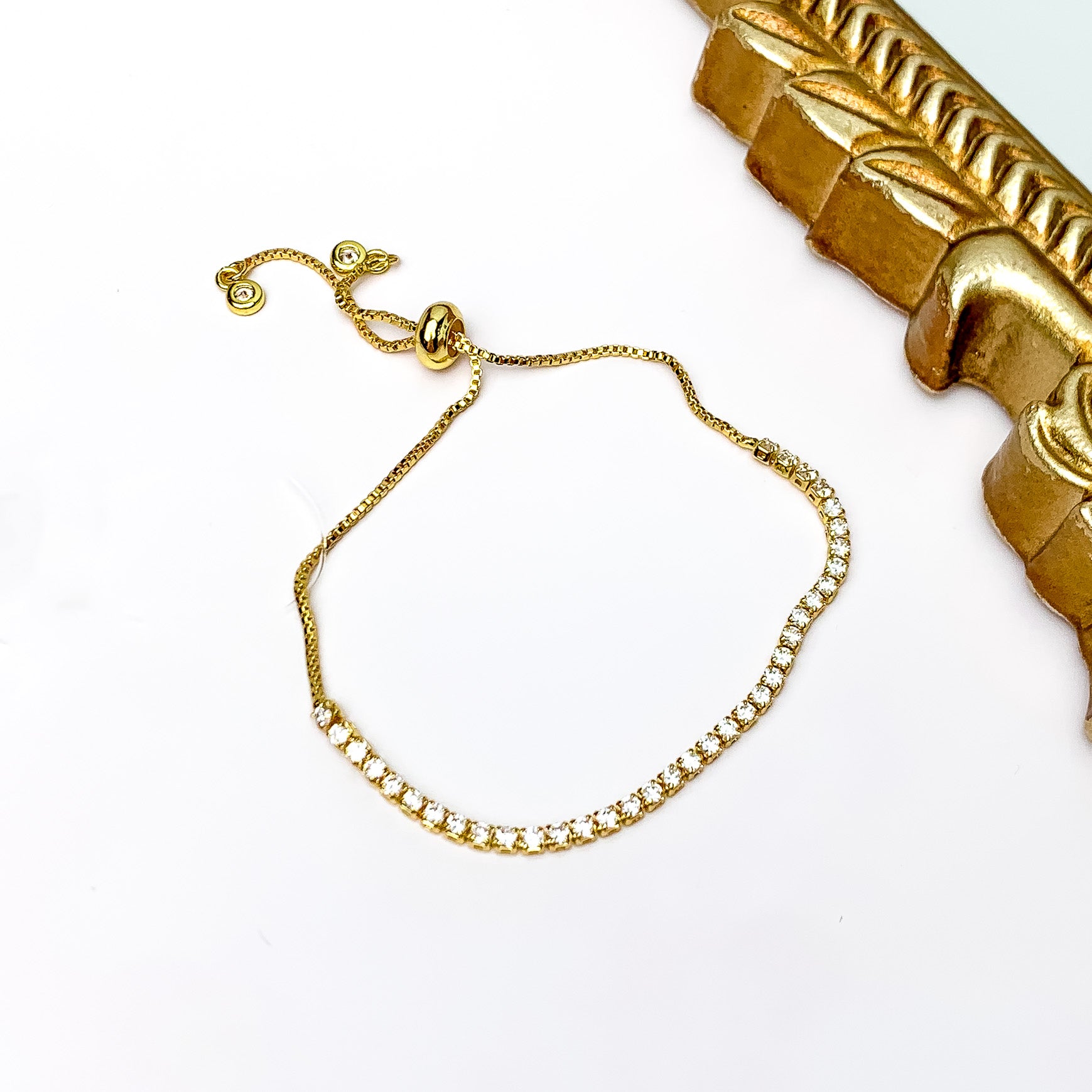 Pictured is a gold chain bracelet with a gold slider bead for adjusting. This bracelet also includes clear crystals on half of the bracelet. This bracelet is pictured on a white background with a gold mirror shown in the top right corner.  