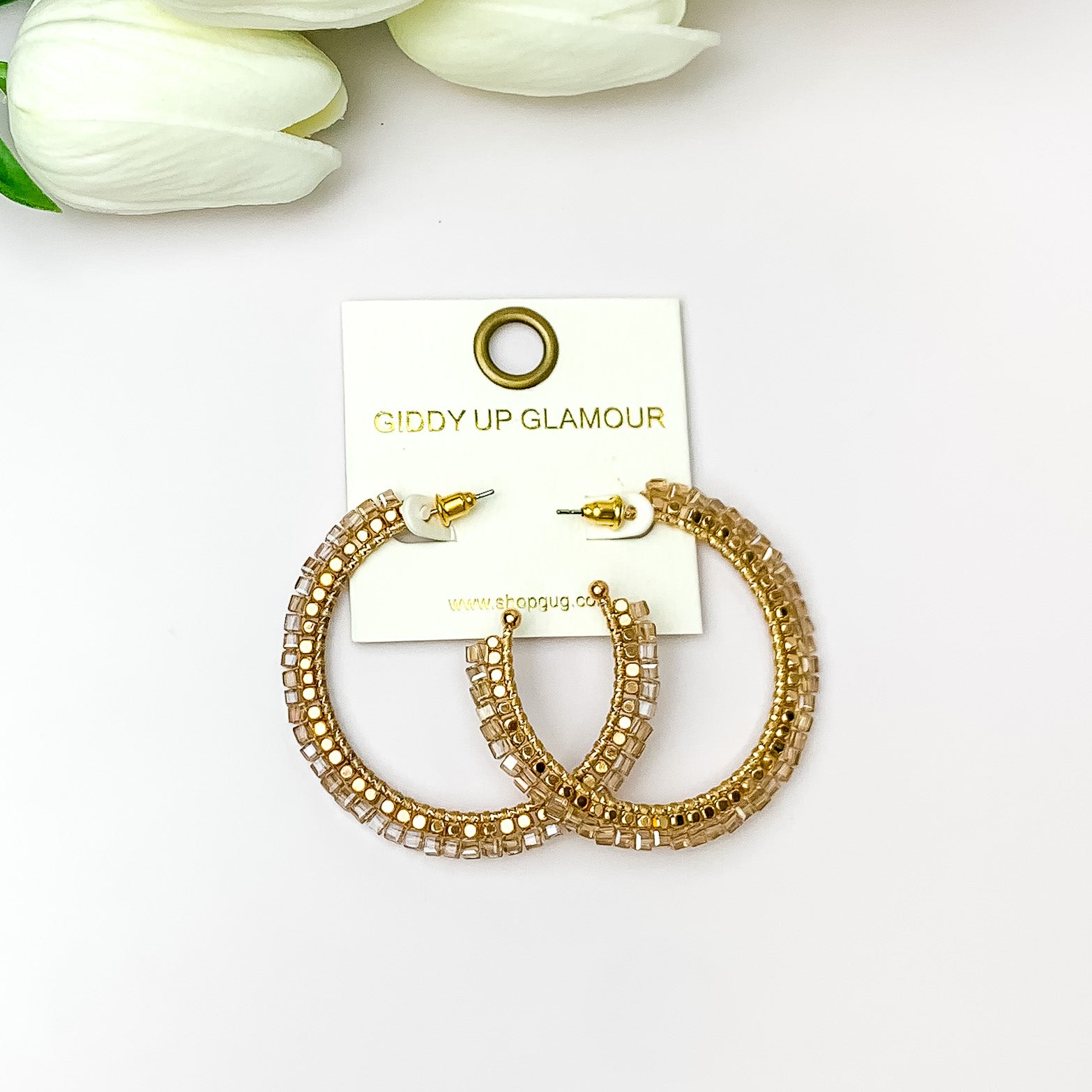 Pictured are circle gold toned hoop earrings with gold beads around it and champagne crystal outline. They are pictured with white flowers on a white background.