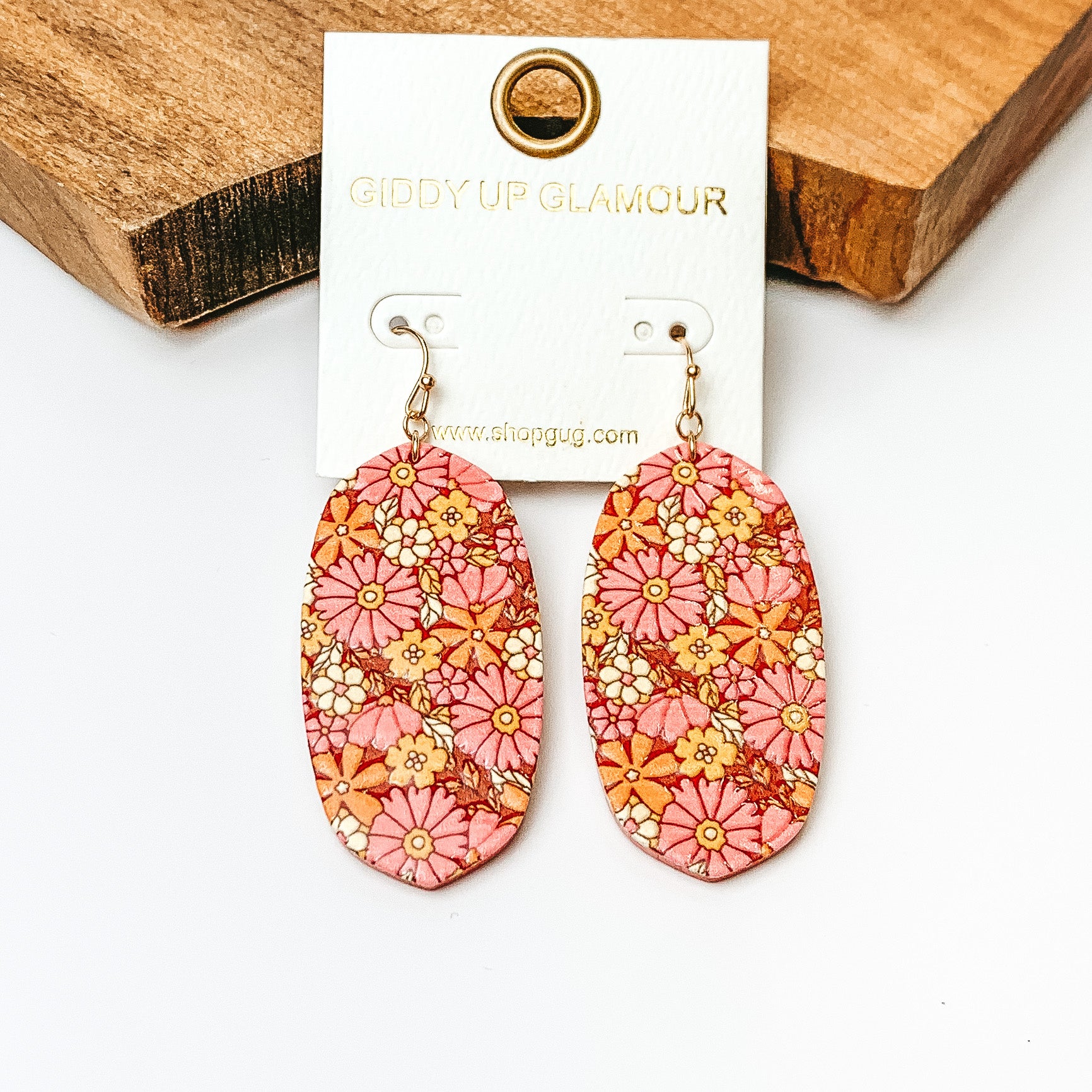 Pictured are oval drop earrings on gold fish hooks. These earrings have floral print in a mix of pinks. These earrings are pictured on a white background in front of a wood block. 