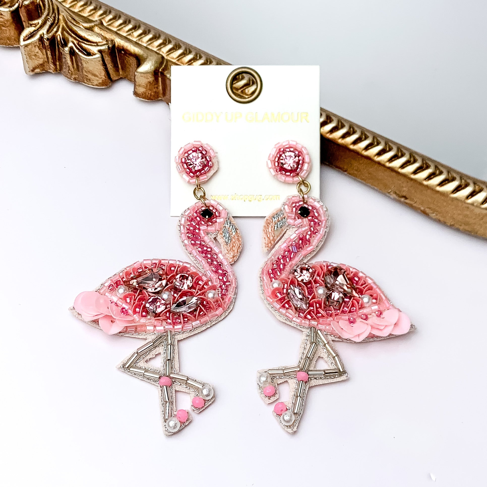 Pictured are beaded flamingo earrings in a light pink beading and different size stones on the body of flamingo. These earrings are propped up on a gold mirror with a white background.