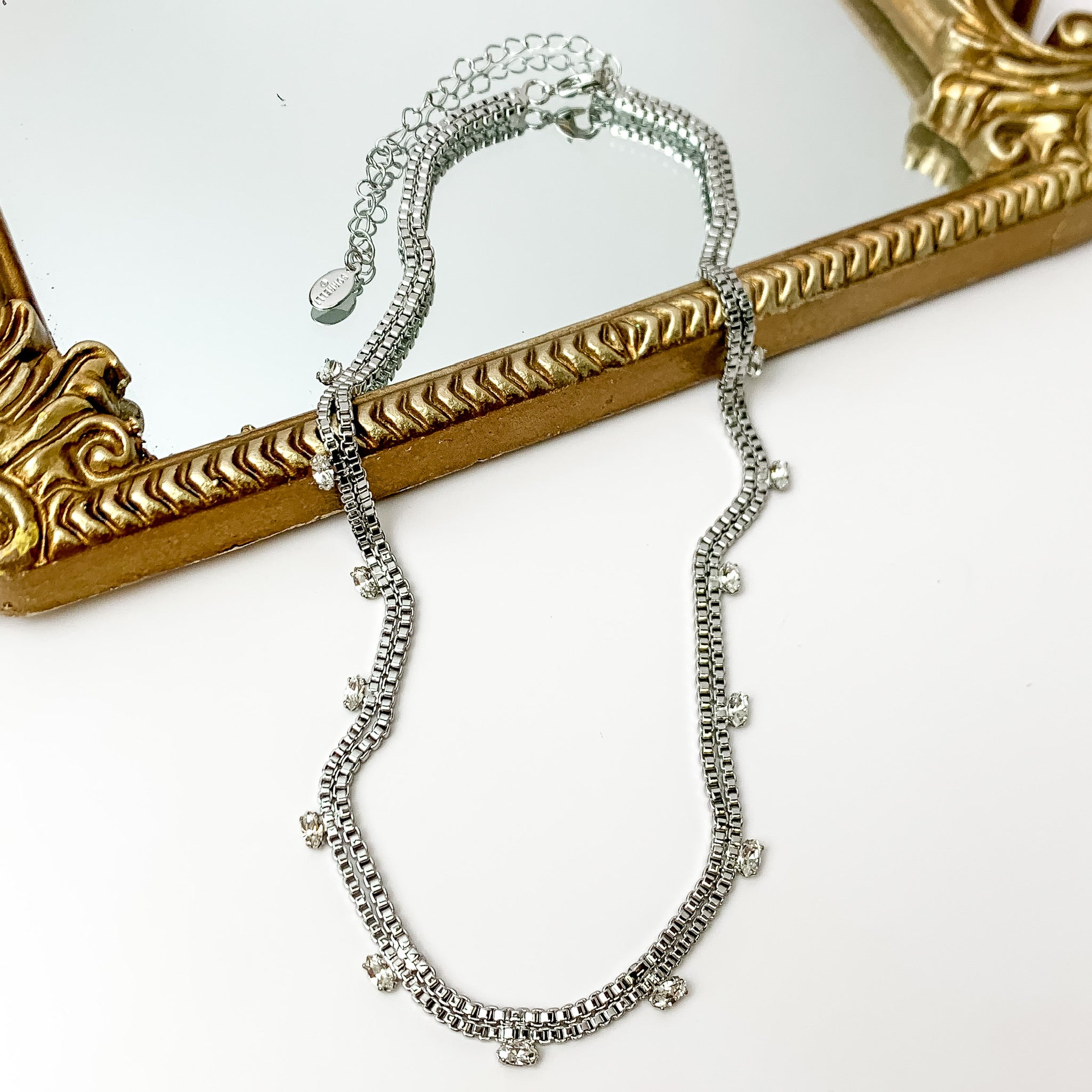Pictured is a double, silver box chain necklace with clear crystal charm. This necklace is pictured partially laying on a gold mirror on a white background.    