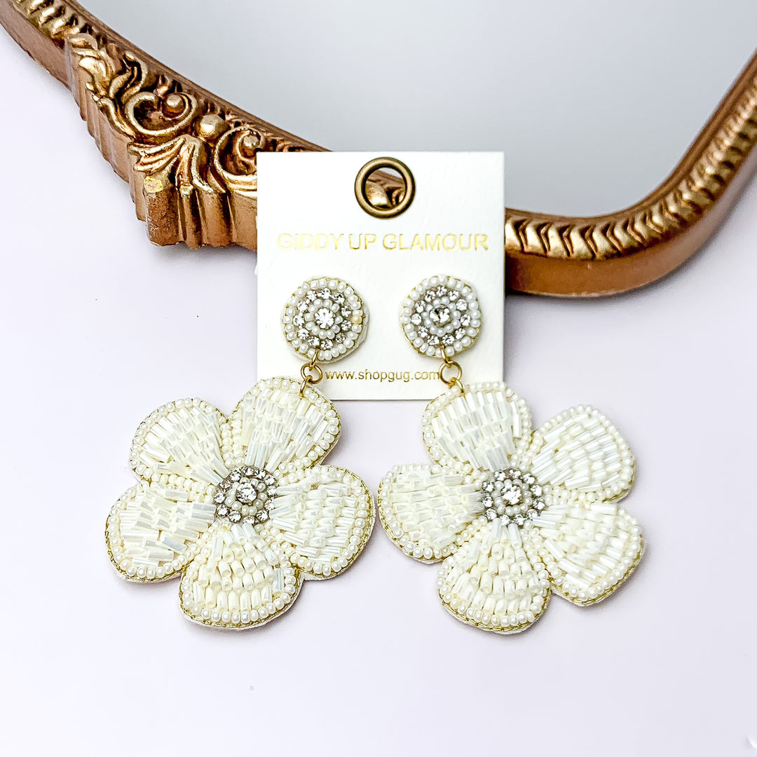  There is hanging beaded flower pendant from the gold stud earrings. The flower pendant is white with gold outline and detailing. These earrings are pictured in front of a gold mirror on a white background.