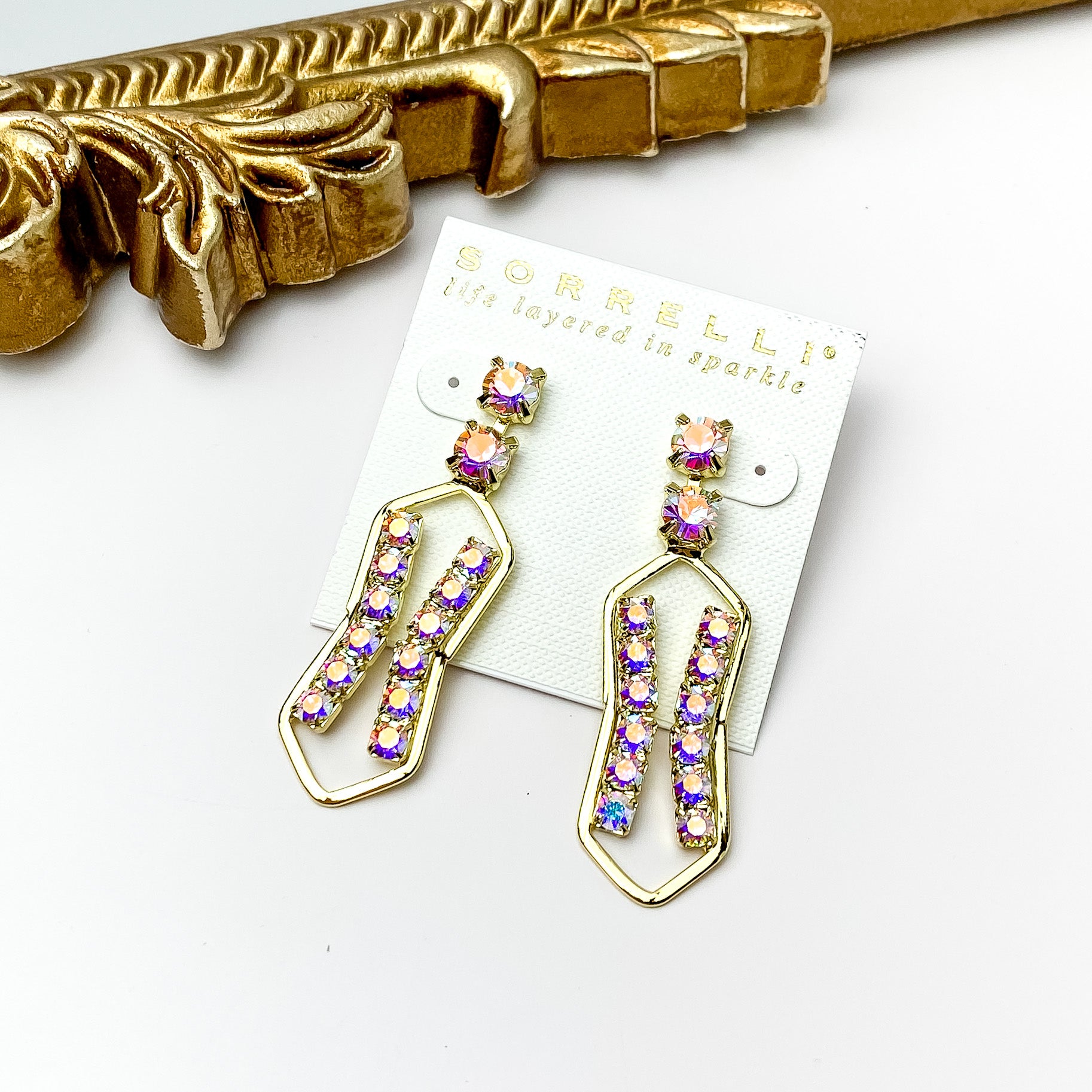 Two ab crystals on a post back with a gold, hourglass shaped drop. The gold drop include a line of ab crystals on each side. These earrings are pictured in front of a gold mirror on a white background. 